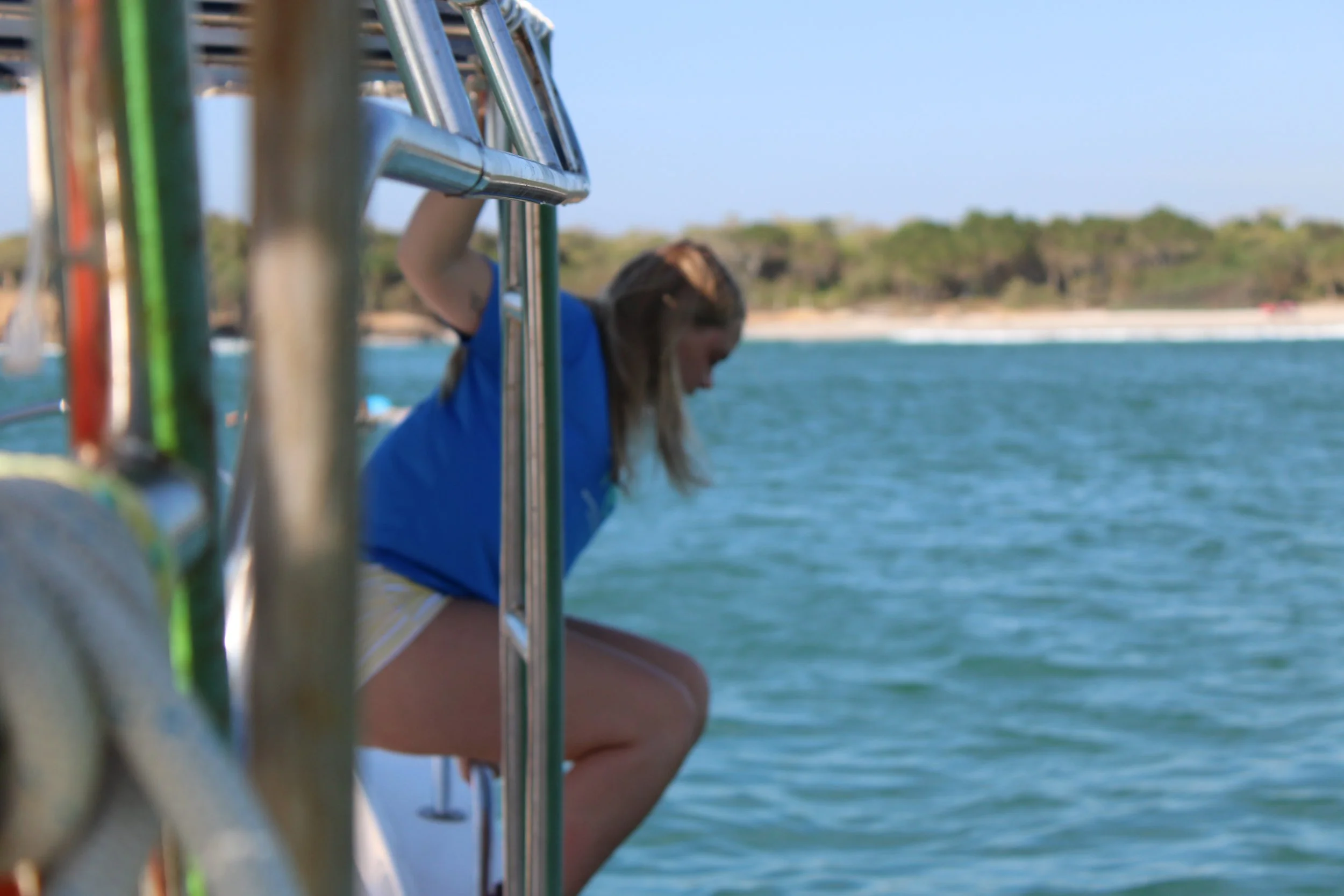 A woman in a blue shirt and striped shorts leaning over the side of a boat near the water, with trees and a sandy shoreline in the background.