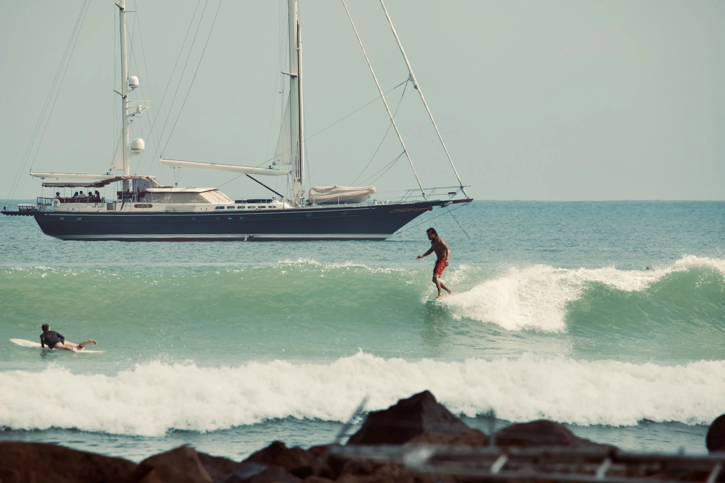 A man surfing on a wave with a large yacht anchored in the background on the ocean.