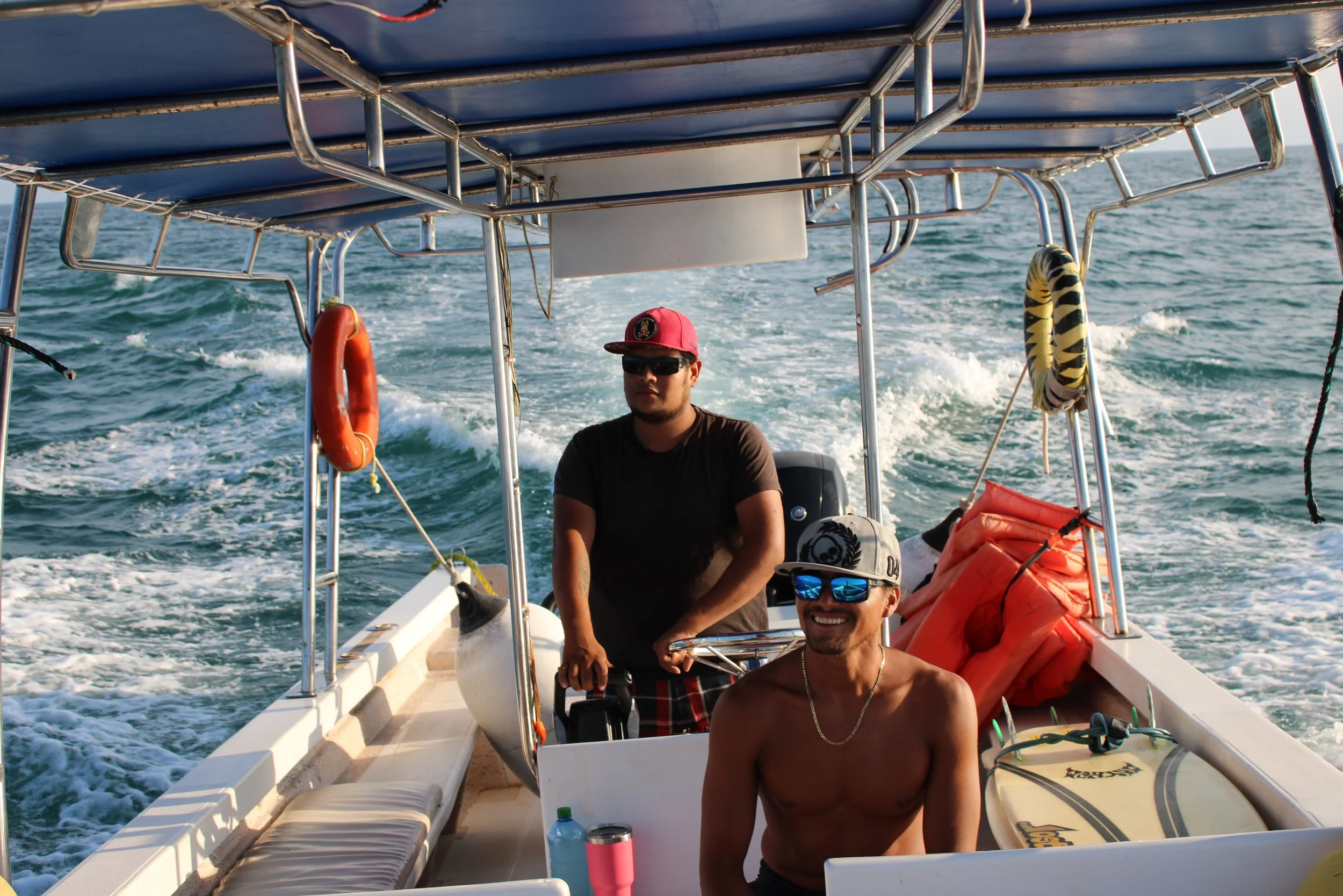 Two men on a boat, one shirtless wearing sunglasses and a cap, the other in a black shirt and red cap, with the ocean and boat wake in the background.