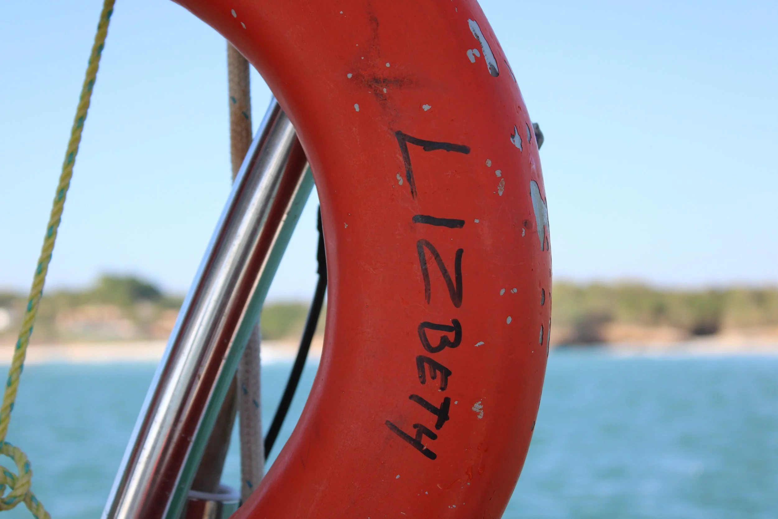Close-up of a red lifebuoy with black writing, hanging on a boat, with water and a distant shoreline in the background.