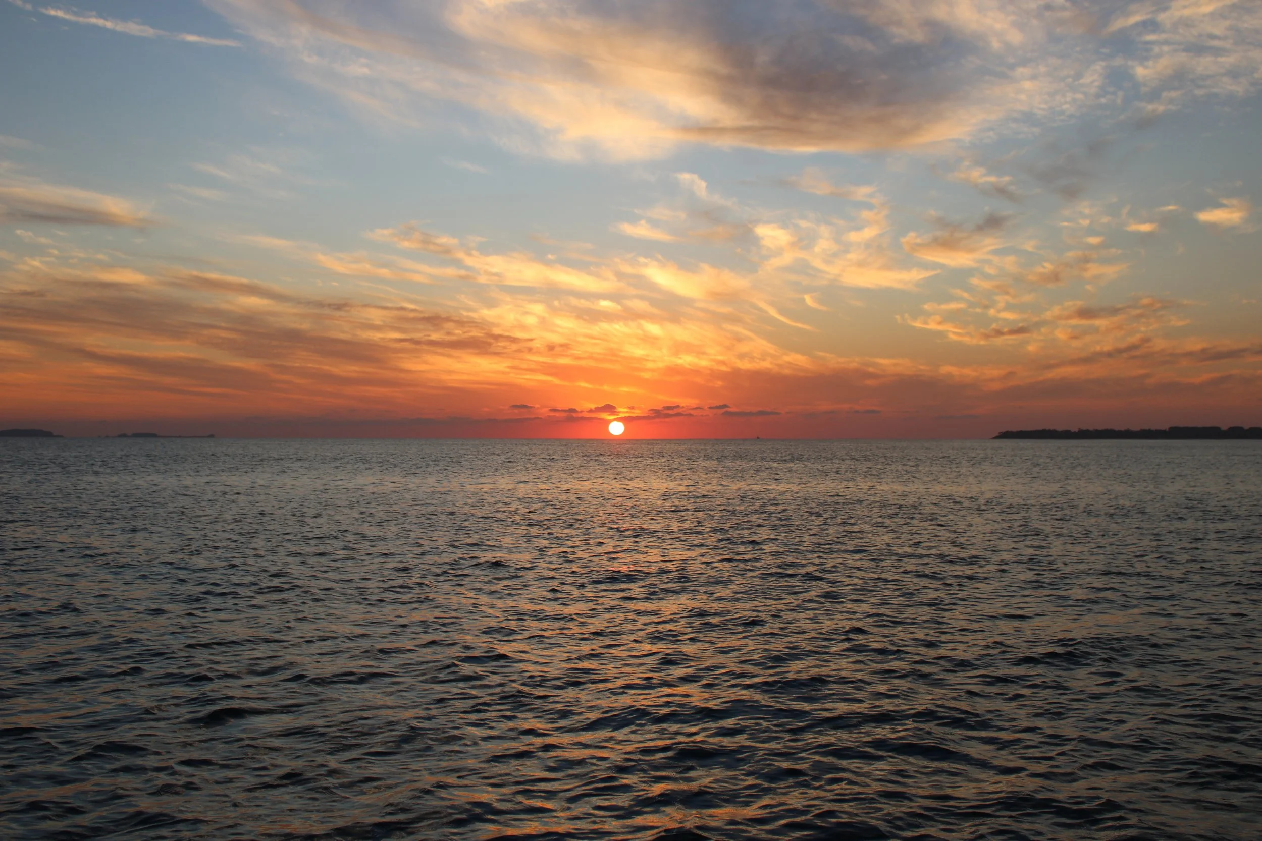 A sunset over the ocean with clouds in the sky and land silhouettes on the horizon.