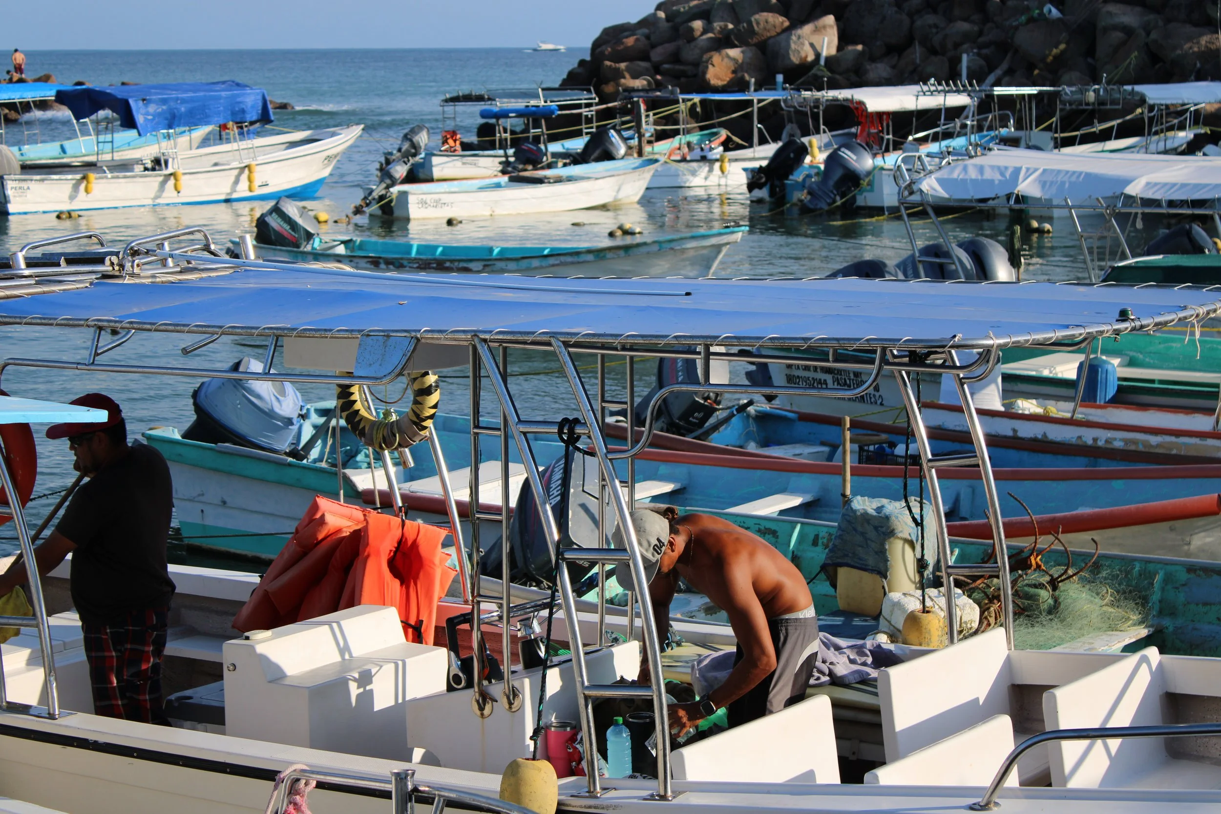 Men working on boats docked at a marina with multiple boats and yachts in the water on a sunny day.