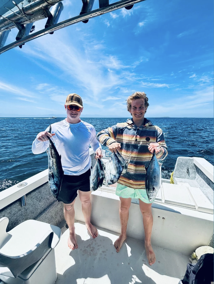 Two men on a boat holding fish they caught, sailing on the ocean under a blue sky with some clouds, smiling at the camera.