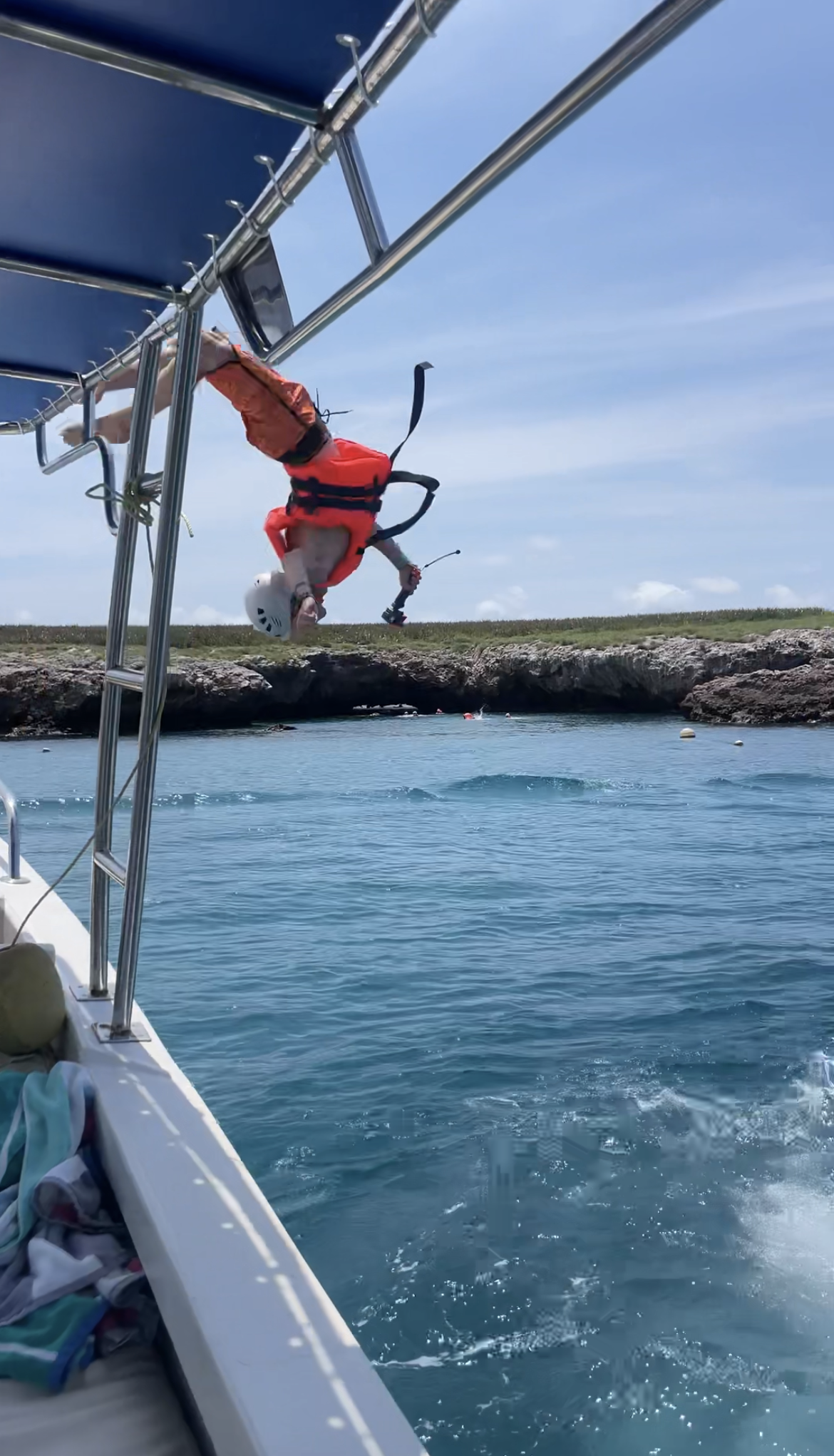 Person in a white helmet and orange life jacket falling off a boat into the water near rocky shoreline.