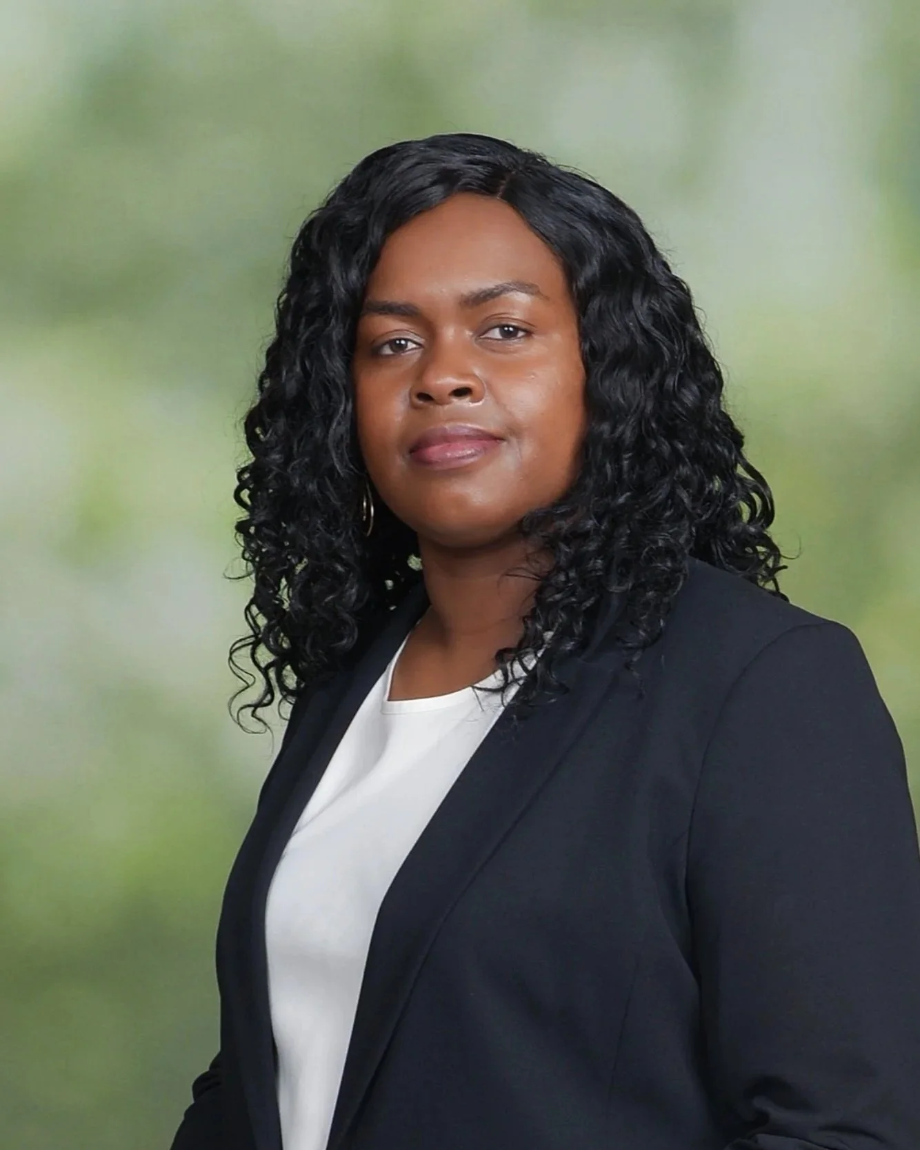 A woman with curly black hair wearing a black blazer over a white top, posing against a blurred green background.