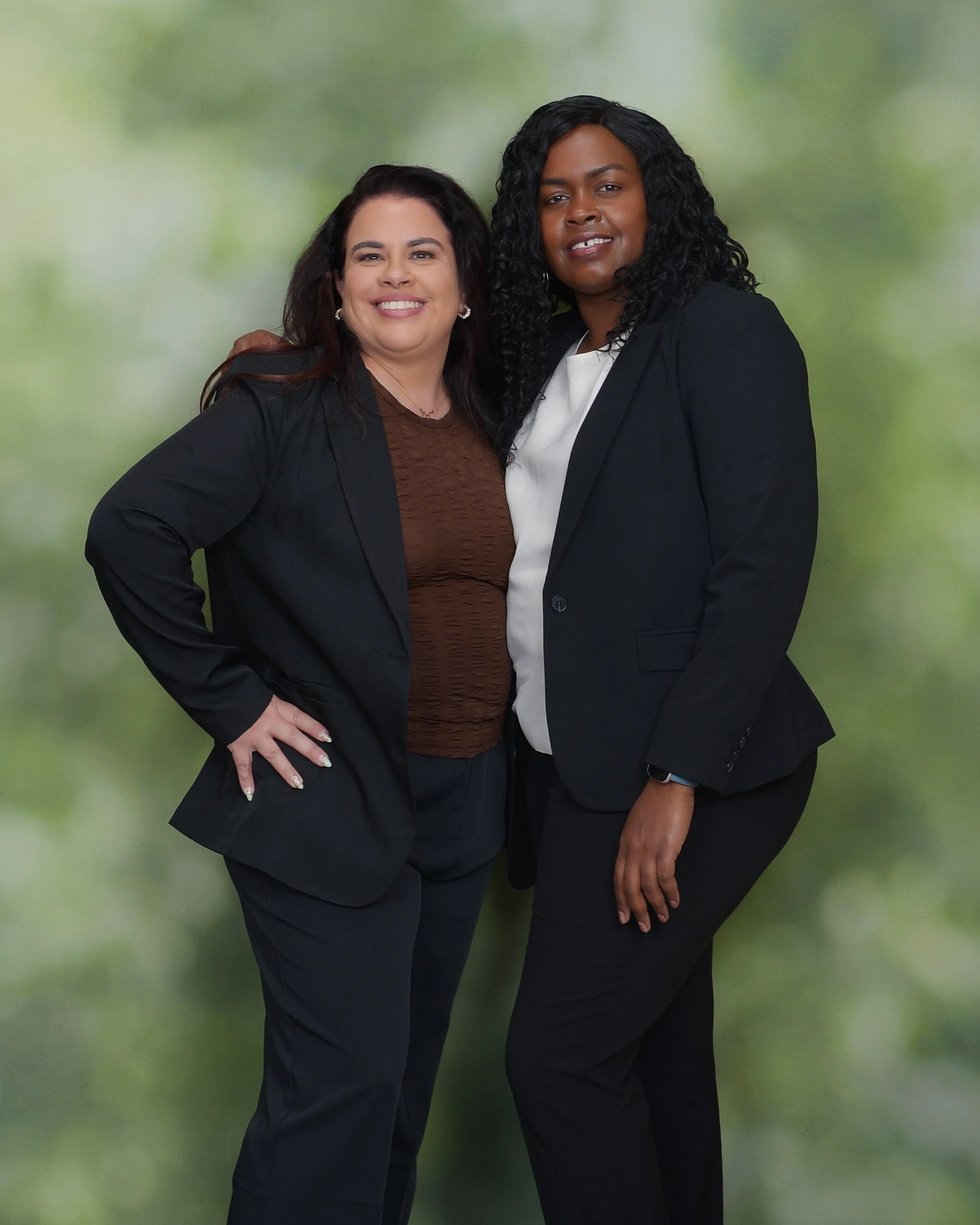 Two women in business attire standing together against a green blurred background, smiling at the camera.
