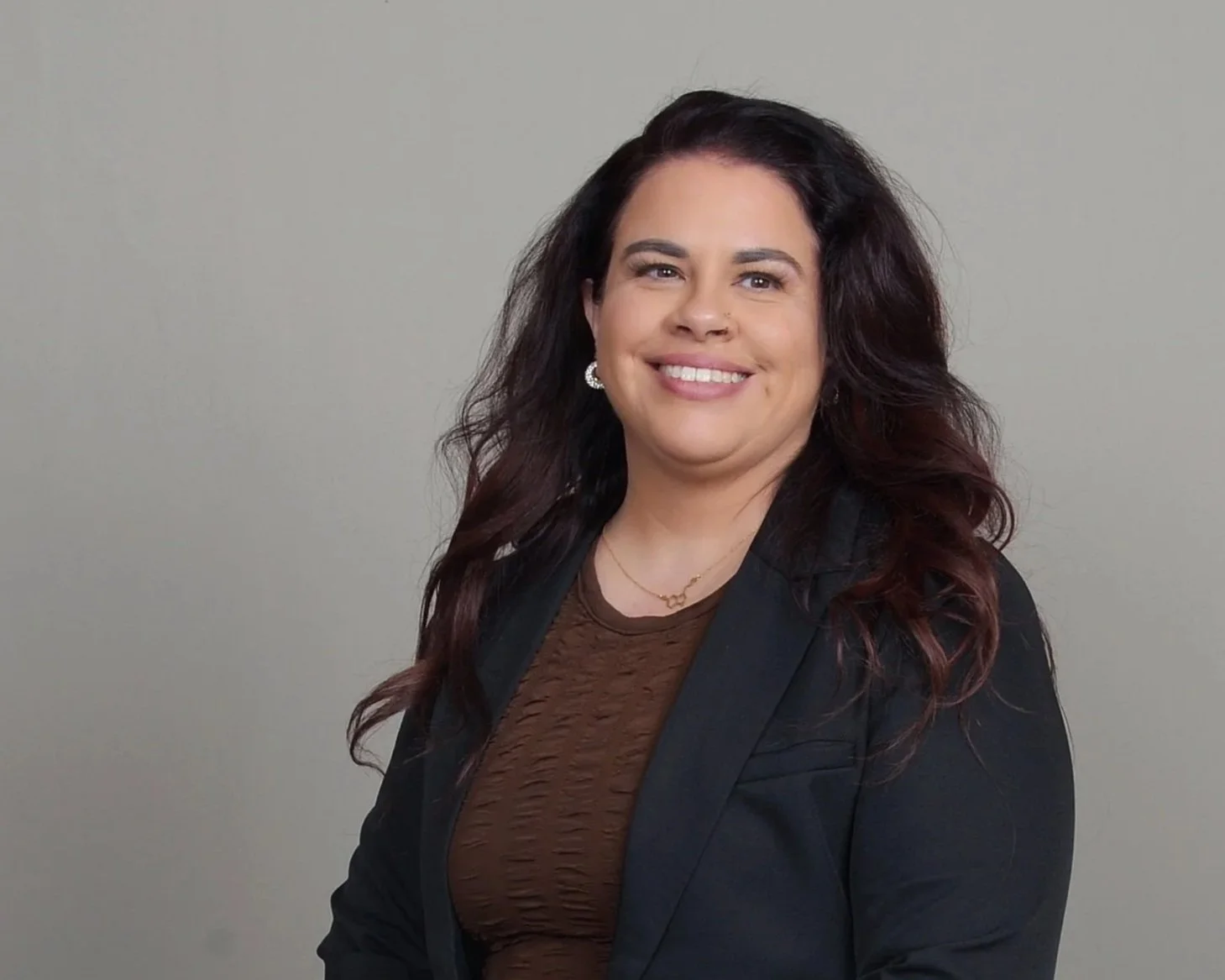 A woman with dark wavy hair smiling, wearing a dark blazer over a brown textured top, earrings, a necklace, and standing against a plain light gray background.