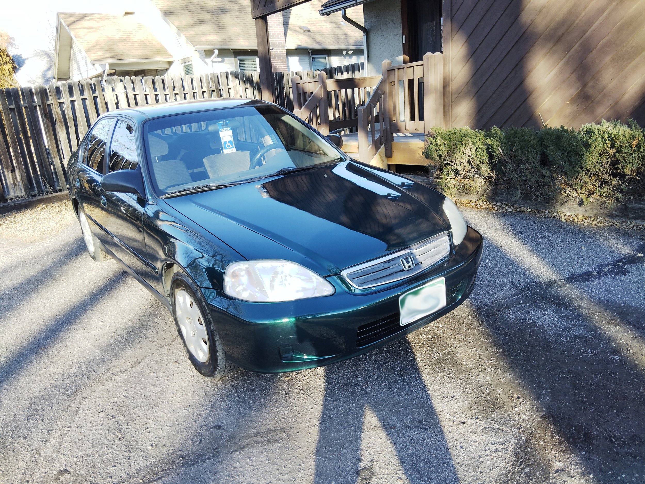 A black Honda Civic parked in a gravel driveway next to a wooden fence and a house with a porch, with shadows cast by nearby trees.