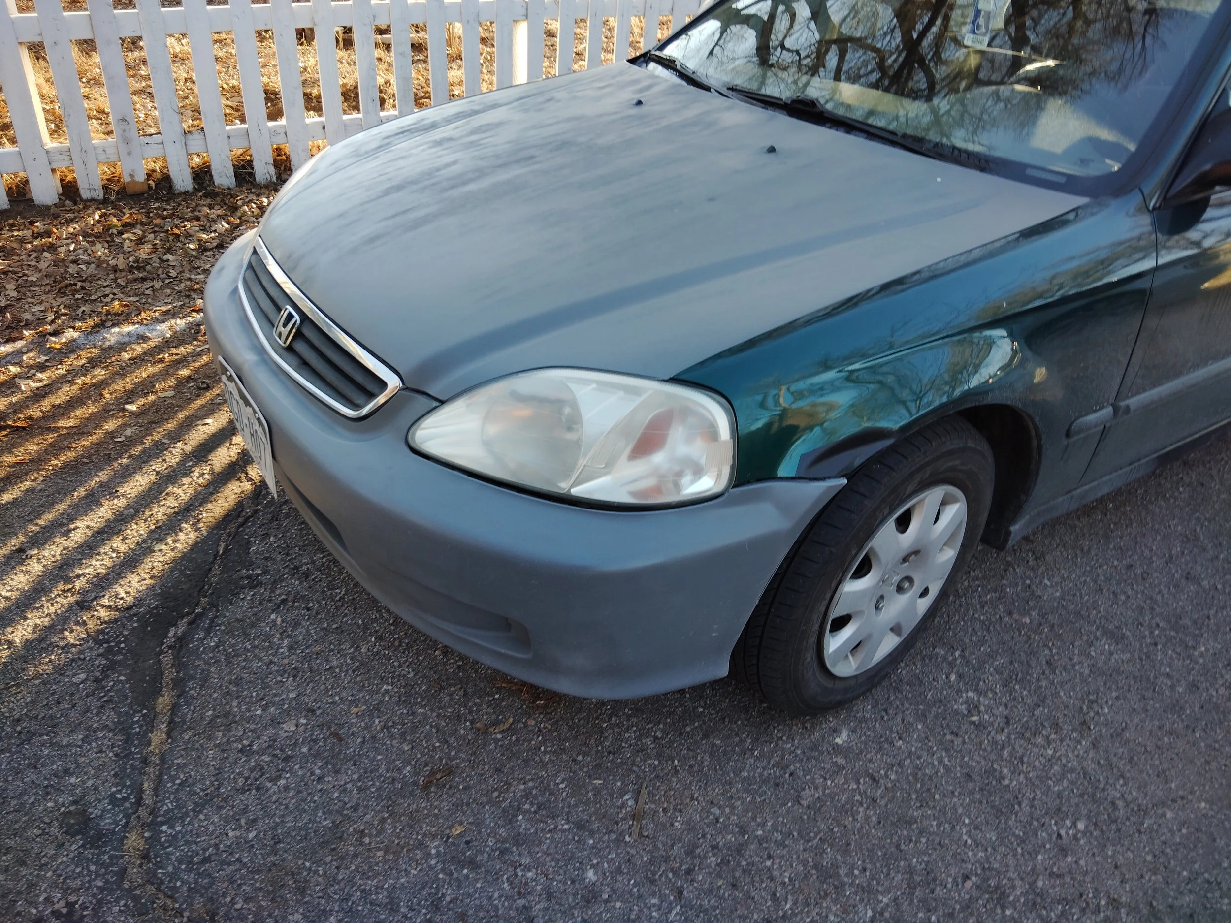 A blue Honda sedan parked on a street beside a white picket fence with fallen leaves in the background.