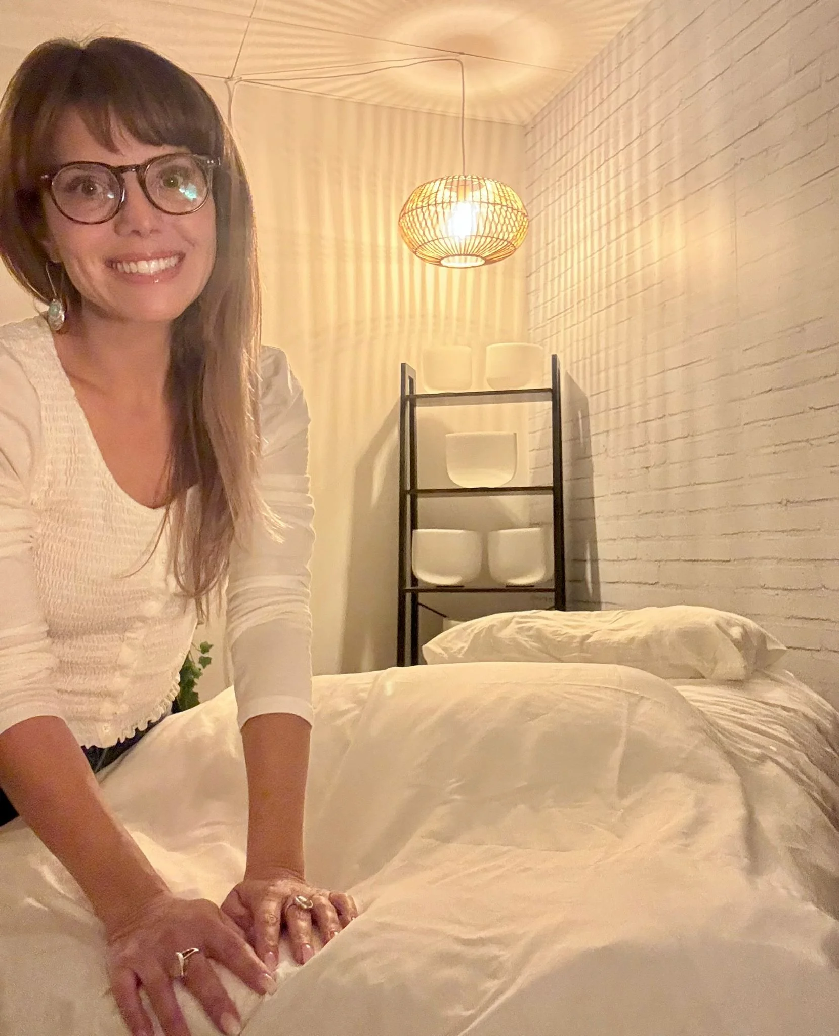 Jennifer Barrow, a woman wearing glasses and a white top, smiling while leaning on a bed with white sheets in a bedroom with a white brick wall and a black shelving unit holding white bowls, illuminated by warm lighting from a woven pendant lamp.