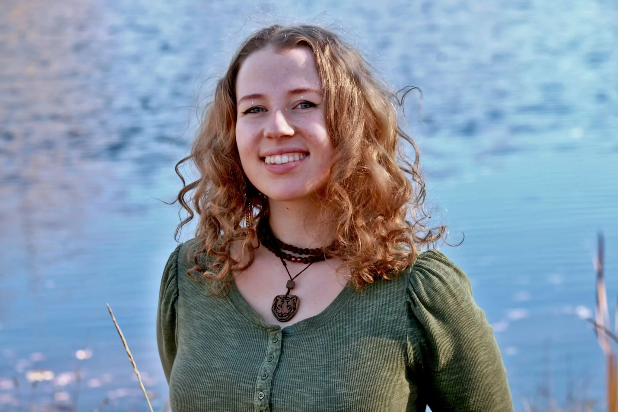 Sarah McDonald, A young woman with curly red hair smiling outdoors with a lake in the background.
