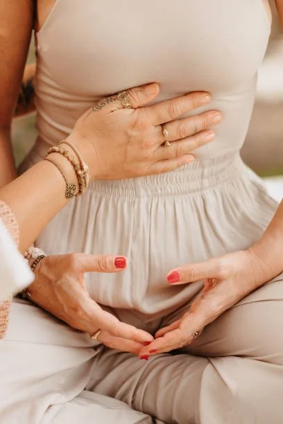Person sitting cross-legged, one with hands forming a heart shape over their belly, with another assisting them in guided breathwork. Both wearing rings and bracelets.