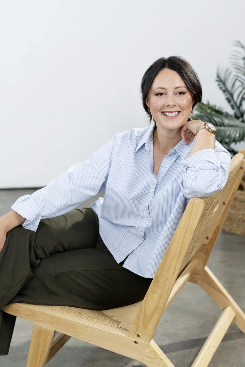 A woman with short dark hair smiling, sitting on a wooden chair, wearing a light blue button-up shirt and dark pants, with a plant in the background.