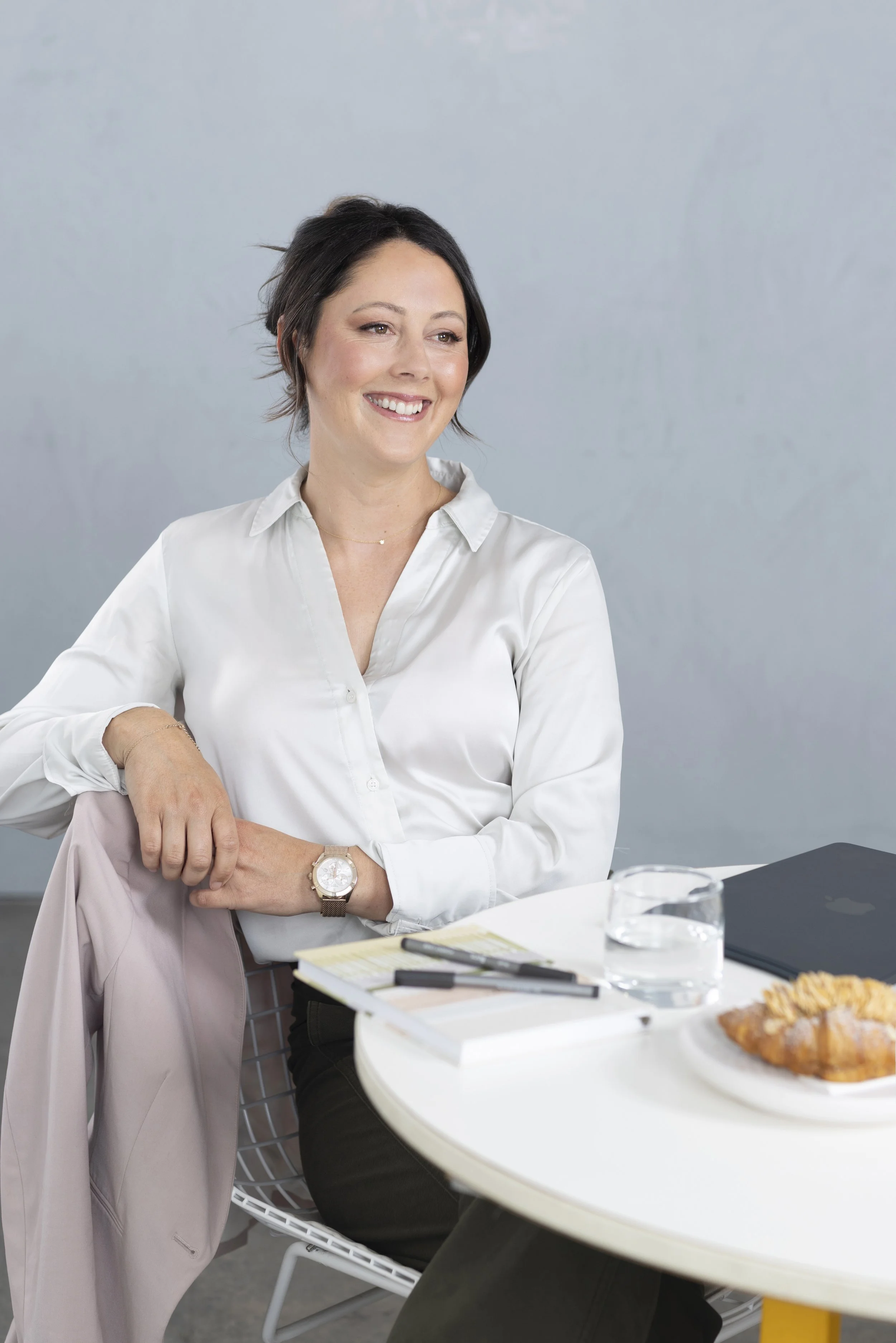 A woman sitting at a table with a pastry, glass of water, notebook, and pen, smiling and looking to her right, wearing a white blouse, gold watch, and a gold necklace. Another person, wearing a light pink blazer, sits beside her with their arm resting on the table.
