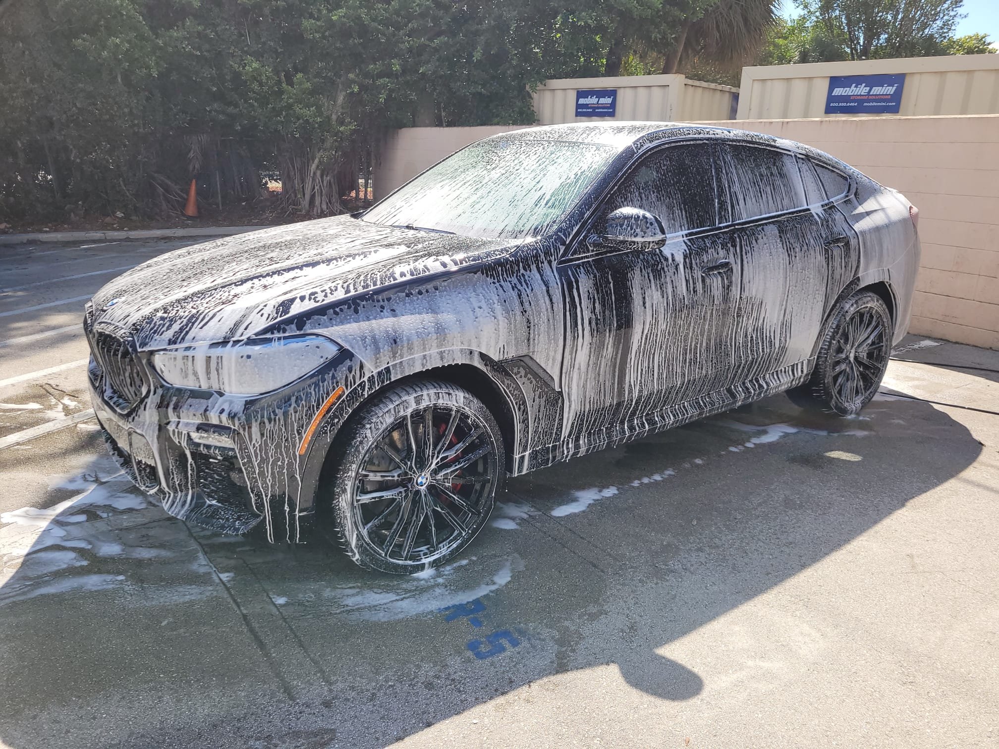 A black BMW sedan covered in white soap suds during a car wash in an outdoor parking lot.
