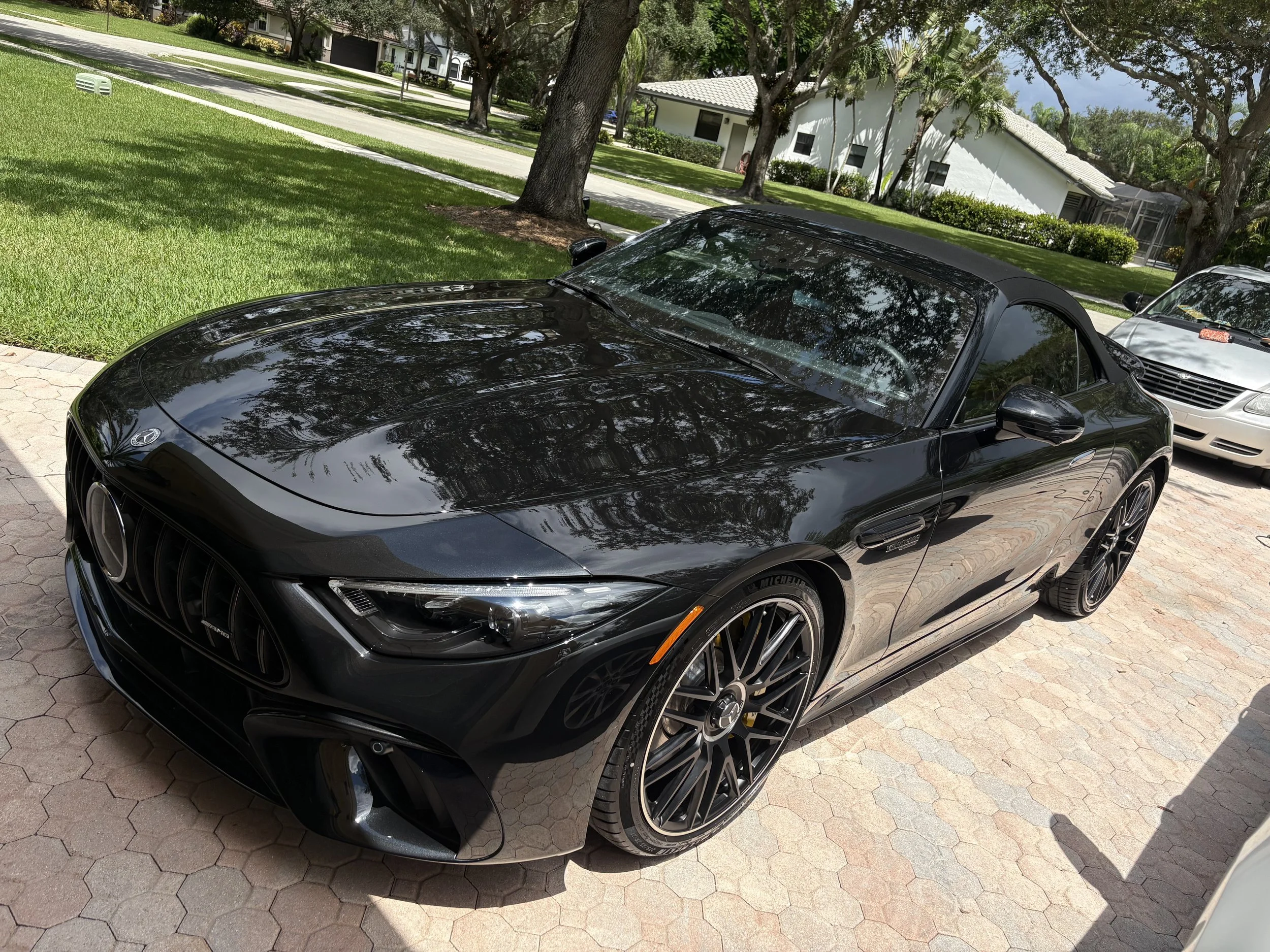 A black convertible sports car parked on a brick driveway with trees and houses in the background.
