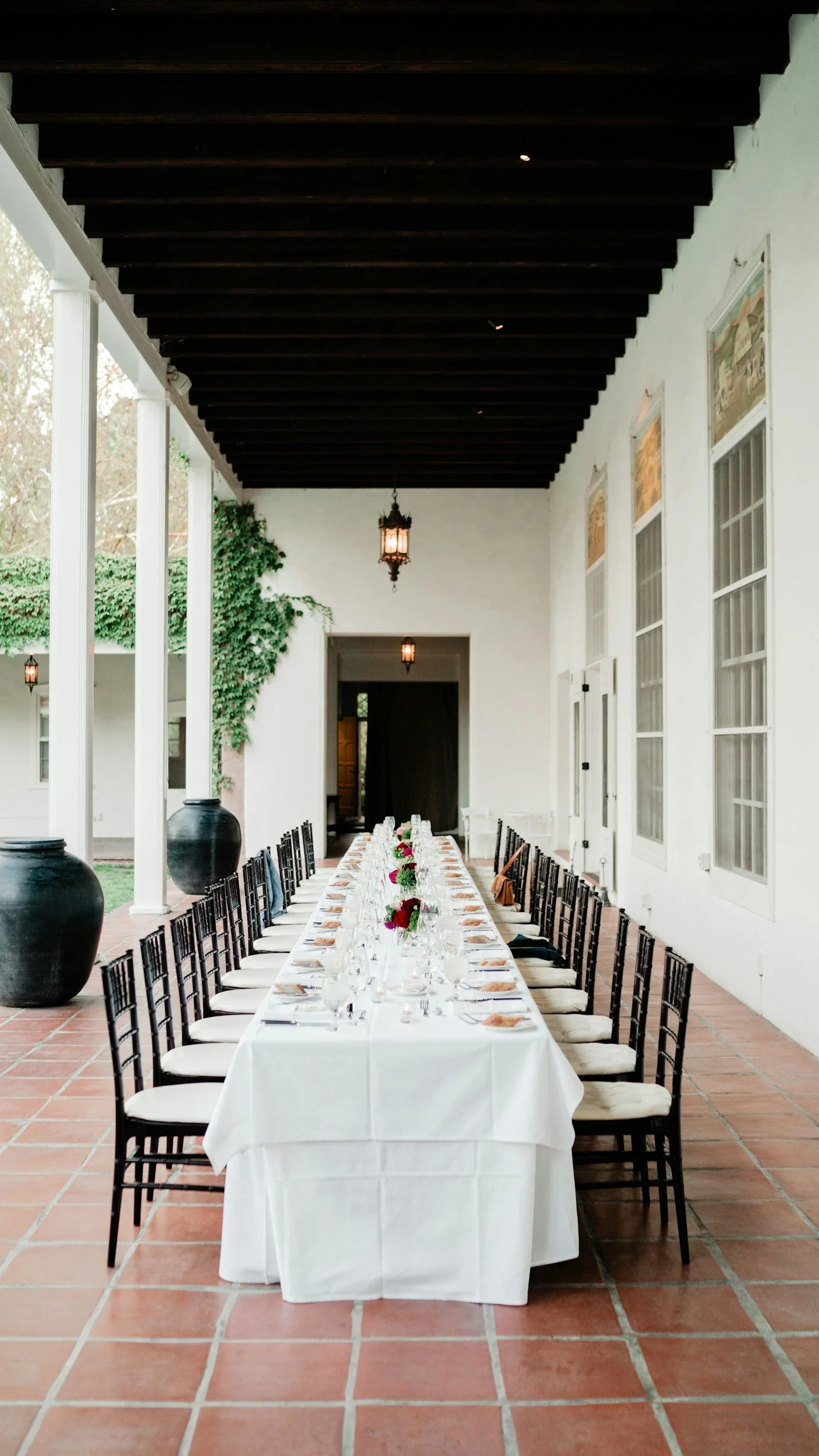 Elegant outdoor dining setup with a long white tablecloth-covered table, set with plates, glasses, cutlery, and small floral centerpieces, under a covered porch with white columns and hanging lanterns, surrounded by white walls, large windows, and terracotta tile flooring.