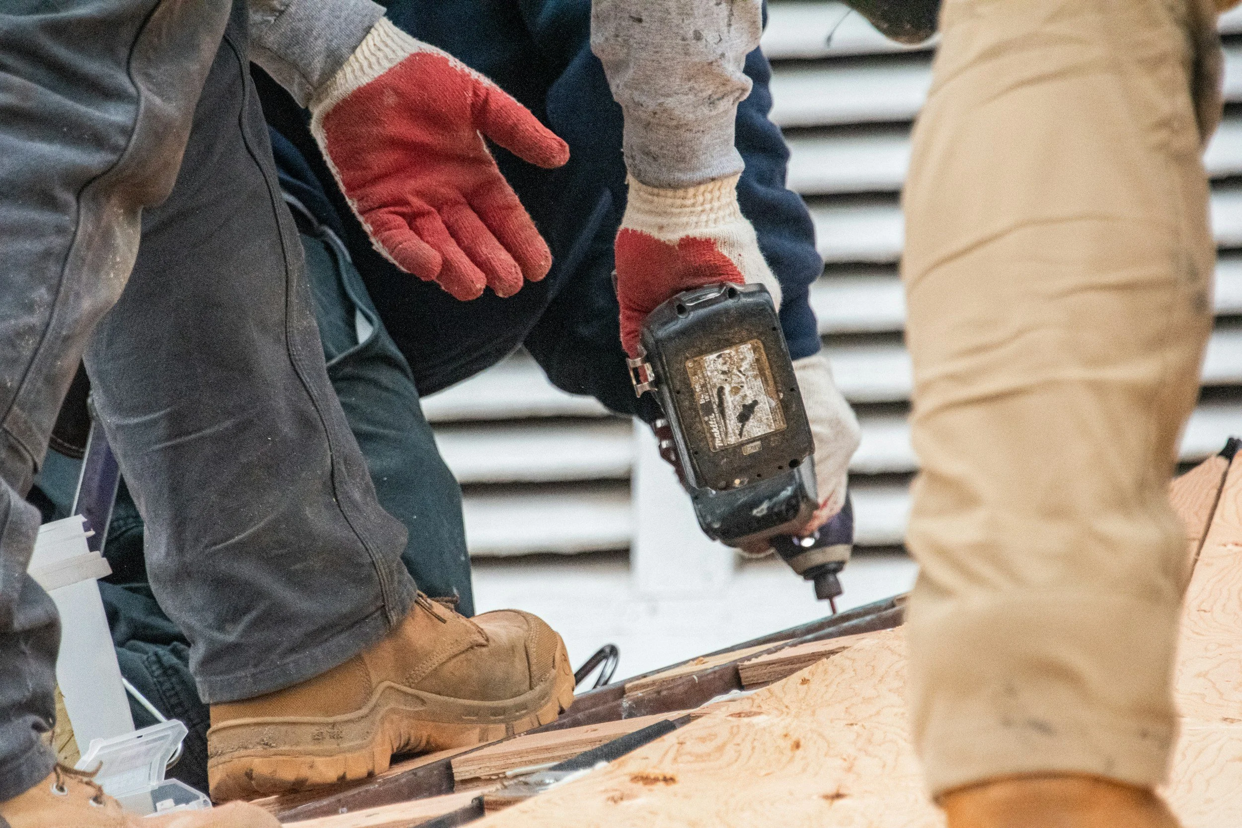 Construction workers installing or working on a wooden structure, with one person using a power drill, wearing gloves, work boots, and protective clothing.