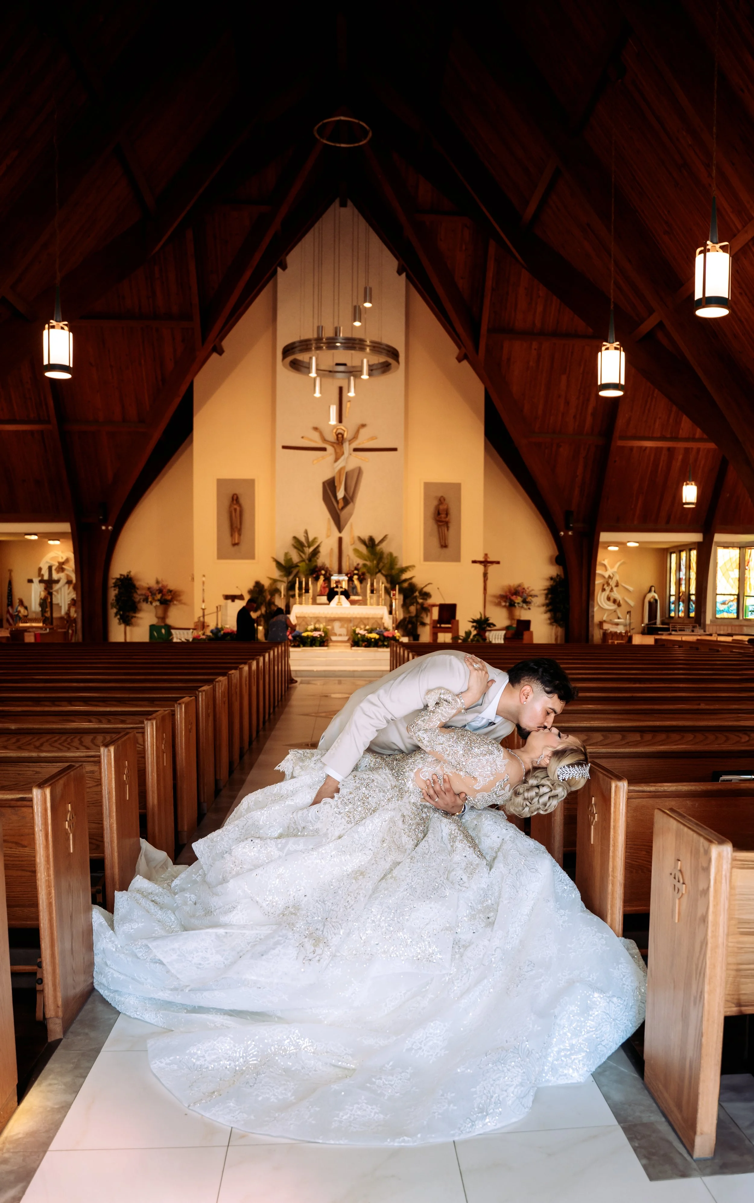 A groom is kissing a bride in a church during their wedding ceremony. The bride is wearing a white wedding gown with lace and sparkles, and the groom is wearing a white suit. They are in the aisle, surrounded by wooden pews and an altar with religious statues and a crucifix in the background.