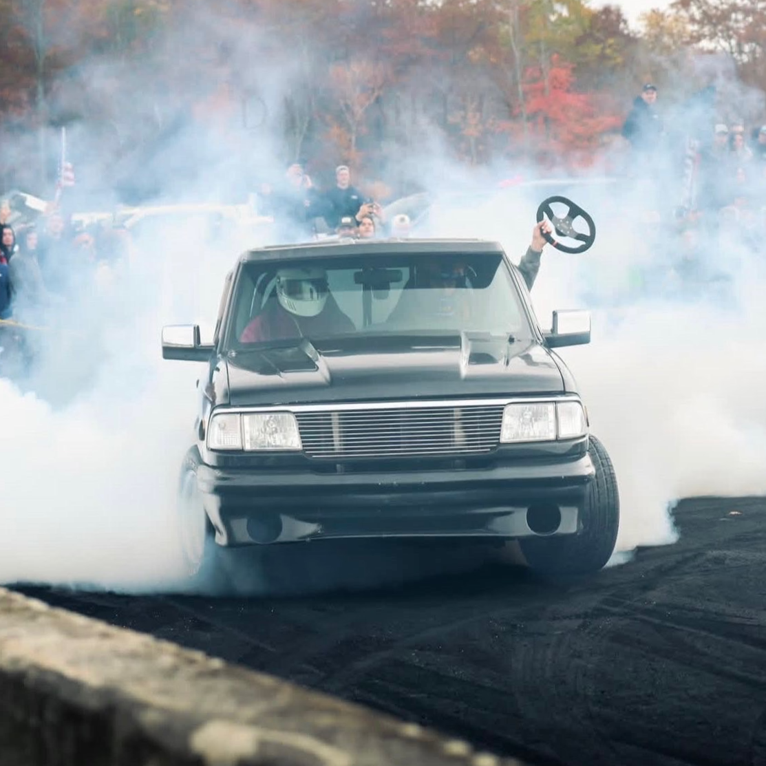 A black race car performs a burnout on a dirt track with smoke billowing around it, while a person holds a steering wheel above the car. Spectators watch in the background, trees with autumn foliage visible.