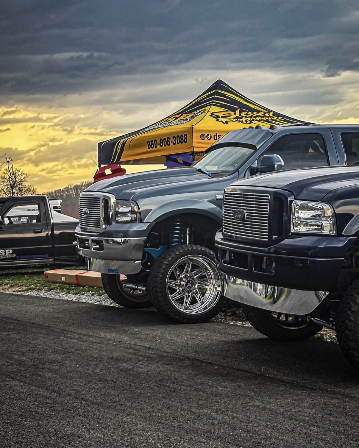 A close-up of a black pickup truck with a chrome grille and shiny rim, parked in front of a yellow and black tent with a phone number. The sky is cloudy with hints of sunlight.