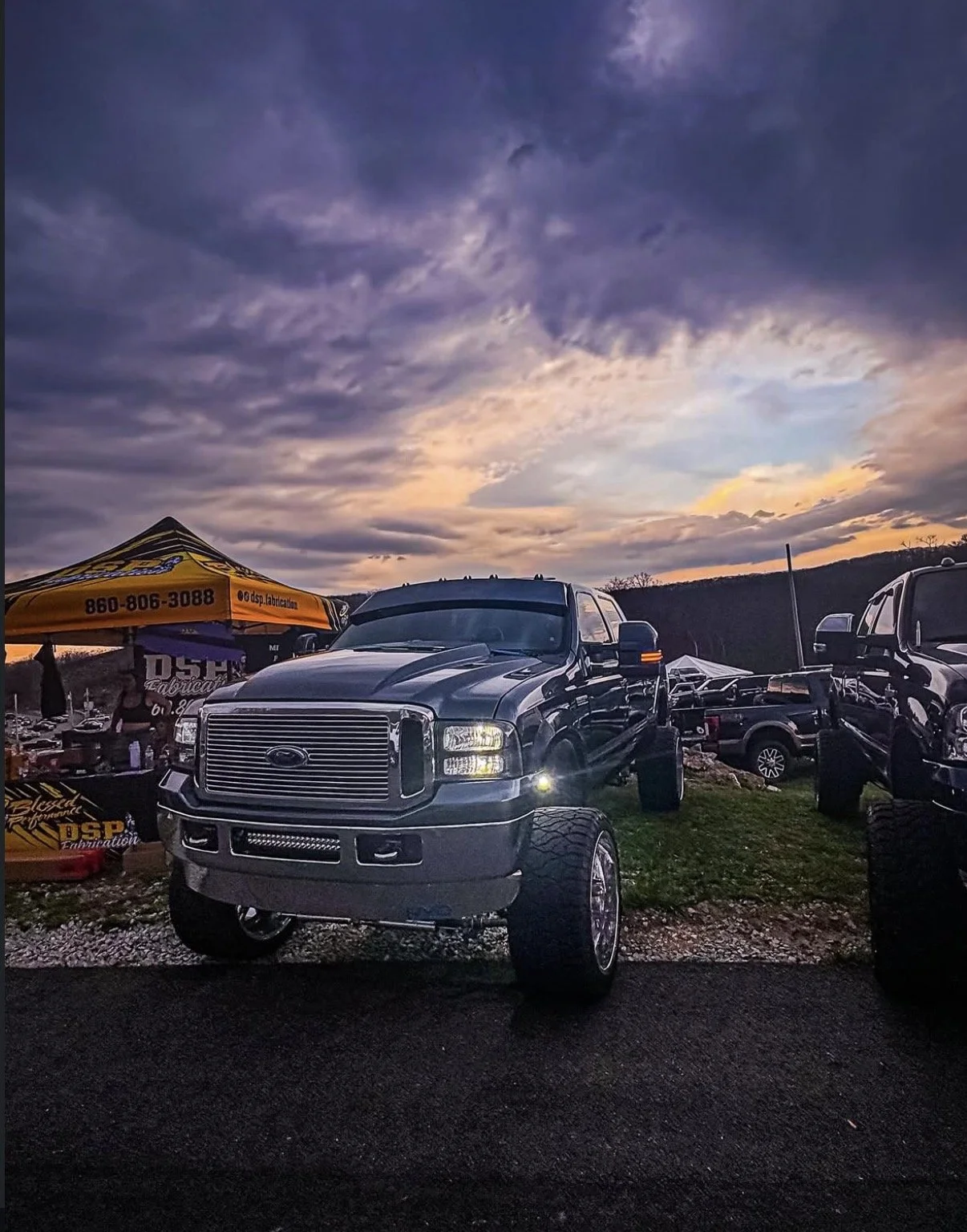 A lifted dark gray or black pickup truck with large wheels parked outdoors at sunset, with other vehicles and a yellow tent in the background.
