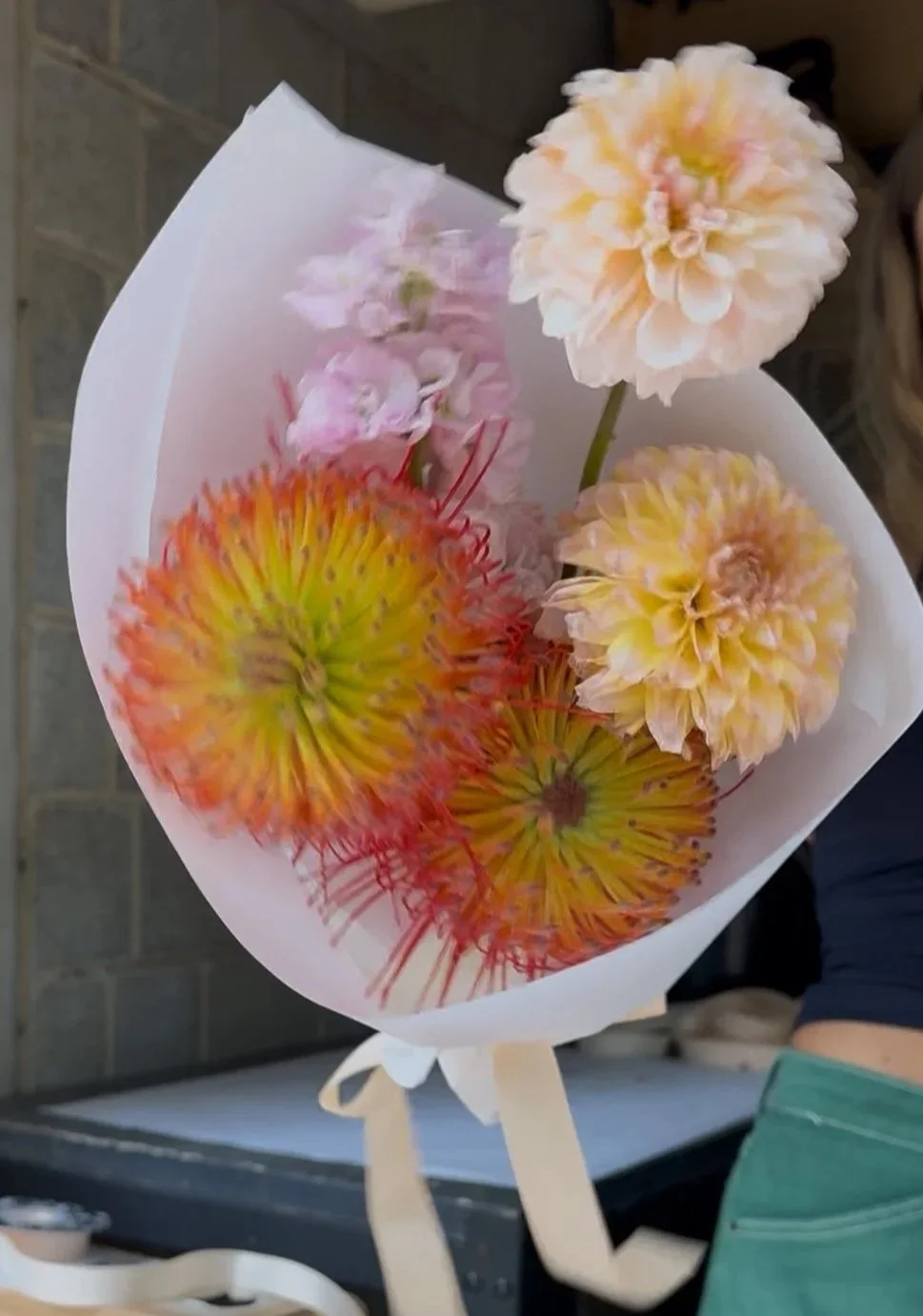 A bouquet of colorful flowers including large cream-colored dahlias, fuzzy orange and yellow pincushion proteas, and pink stock, wrapped in white paper. Florist showcasing a "make your own bouquet" design from interactive flower bar