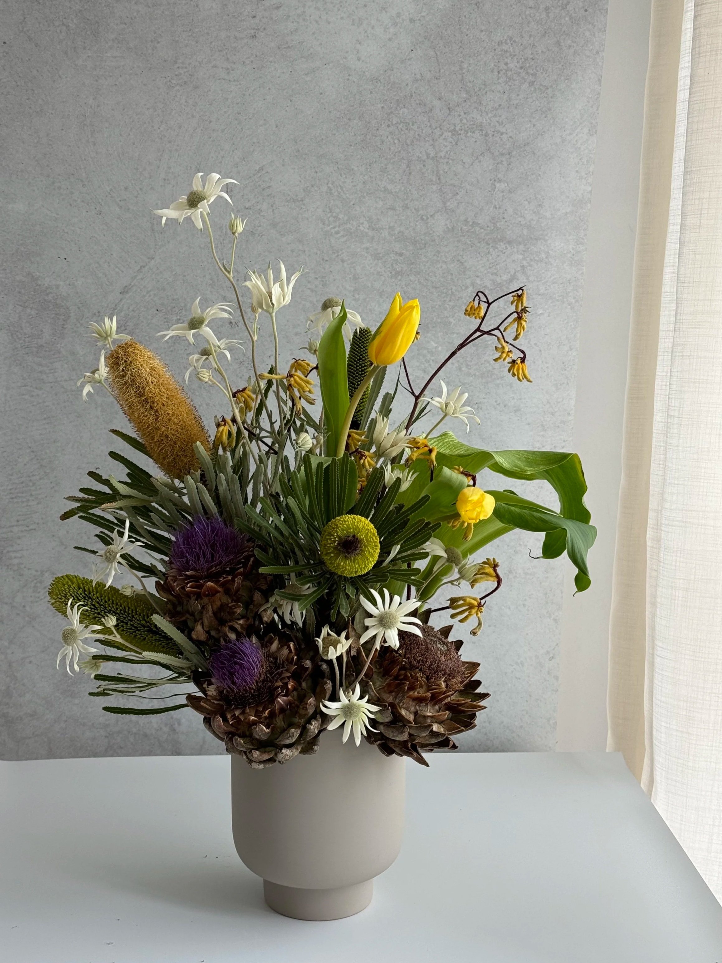 A floral arrangement in a grey vase with yellow, white, purple, and green flowers, set against a light grey wall, designed for a business flower subscription service