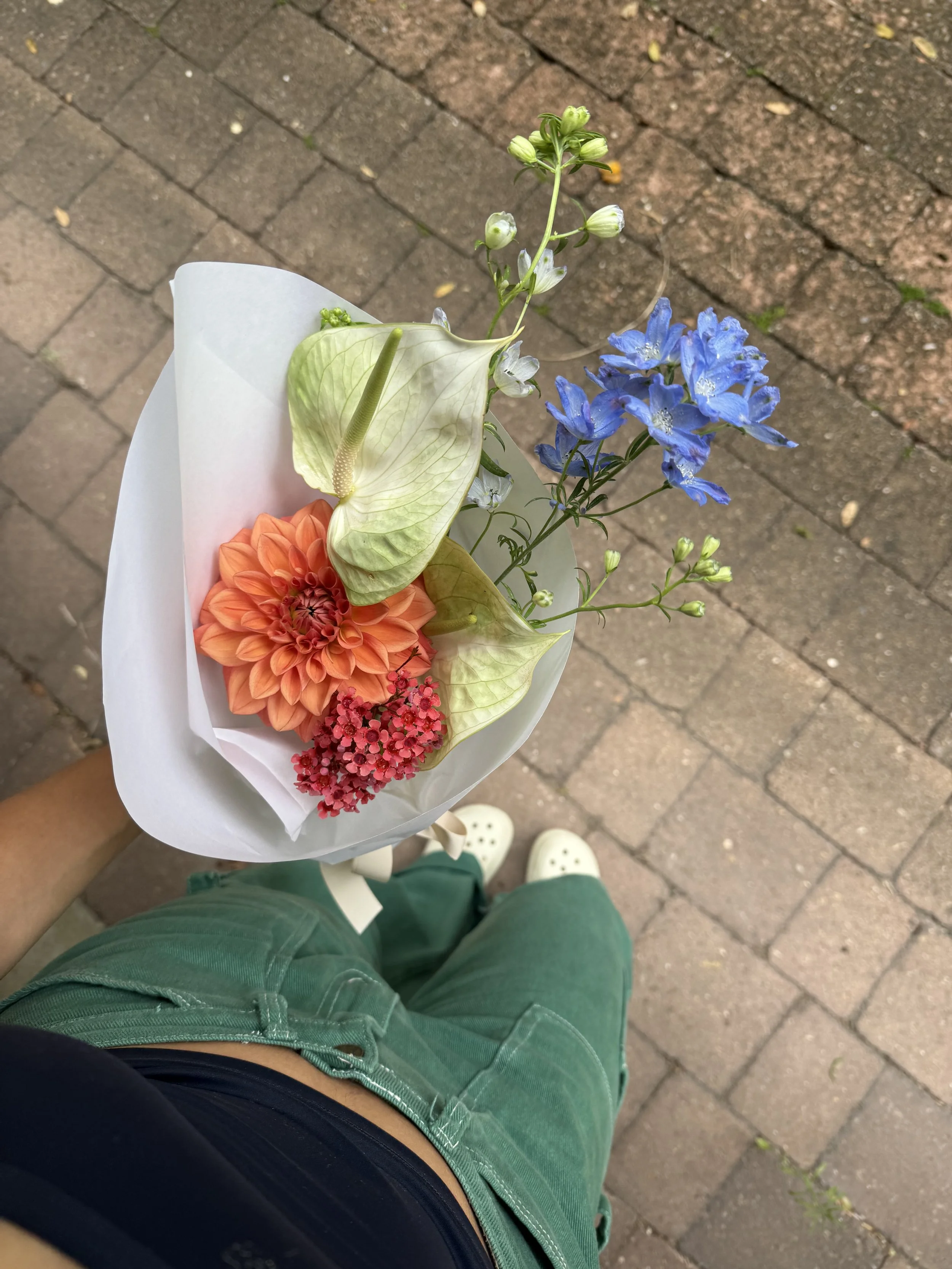 Florist holding a bouquet of multicolored flowers, including an orange dahlia, a white anthurium, blue delphiniums, showcasing what bouquets you can make with Lilac Lane interactive flower bar.