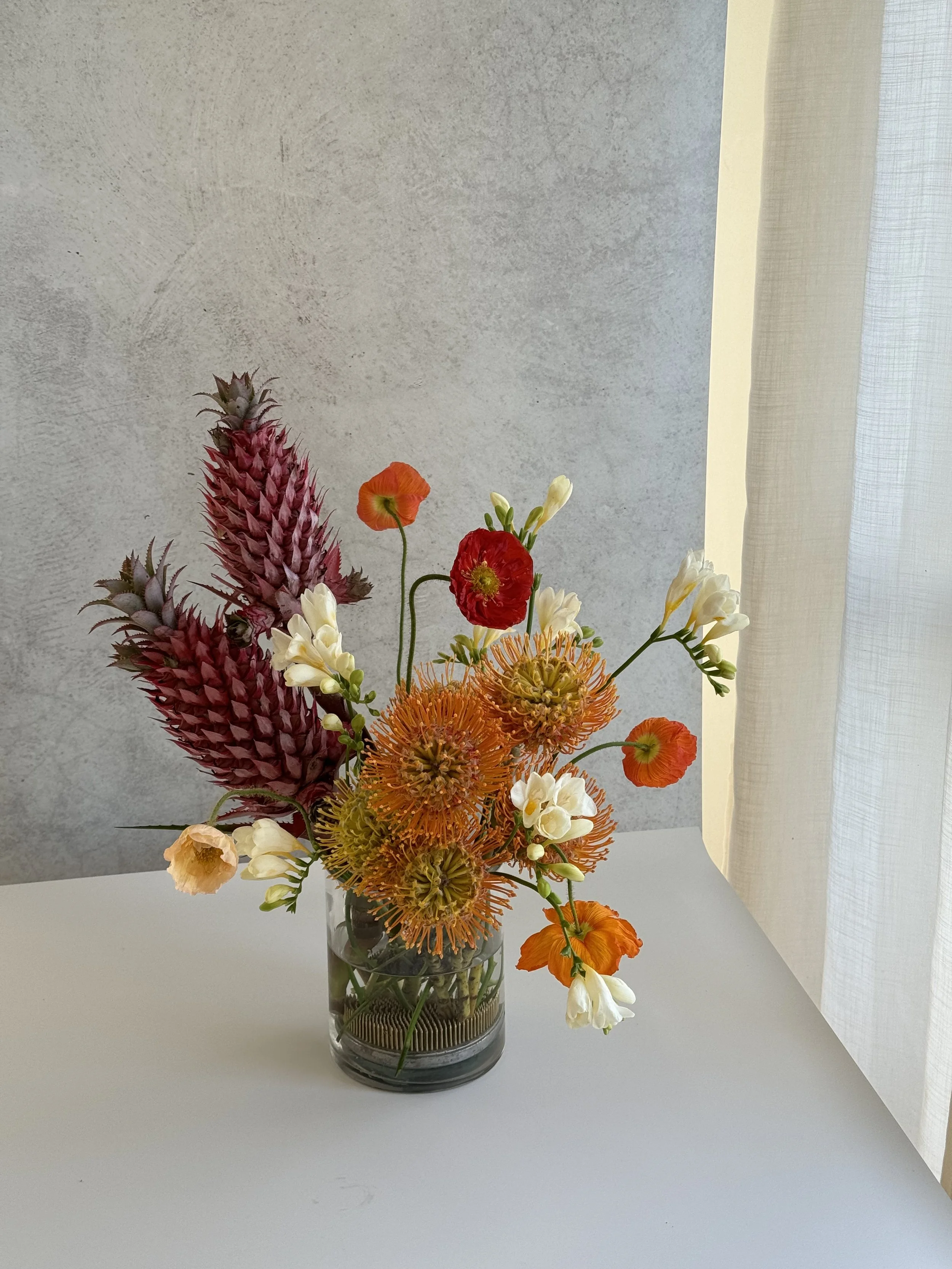 red, orange, white flower arrangement in a glass vase on a white surface. poppies, pineapples and pincushions 