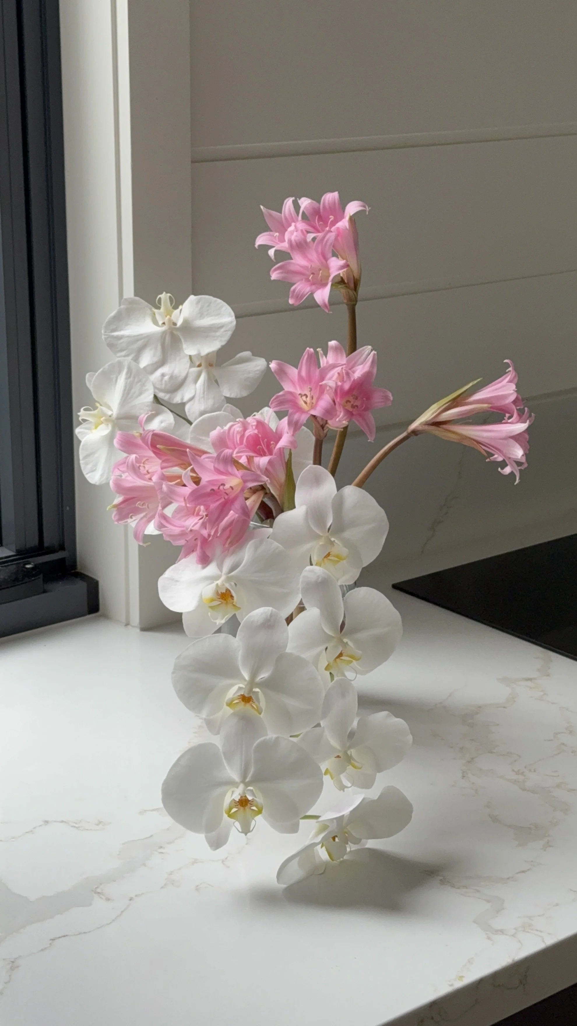 A small floral design of white orchids and pink belladonna lilies on a white marble surface styling a kitchen bench