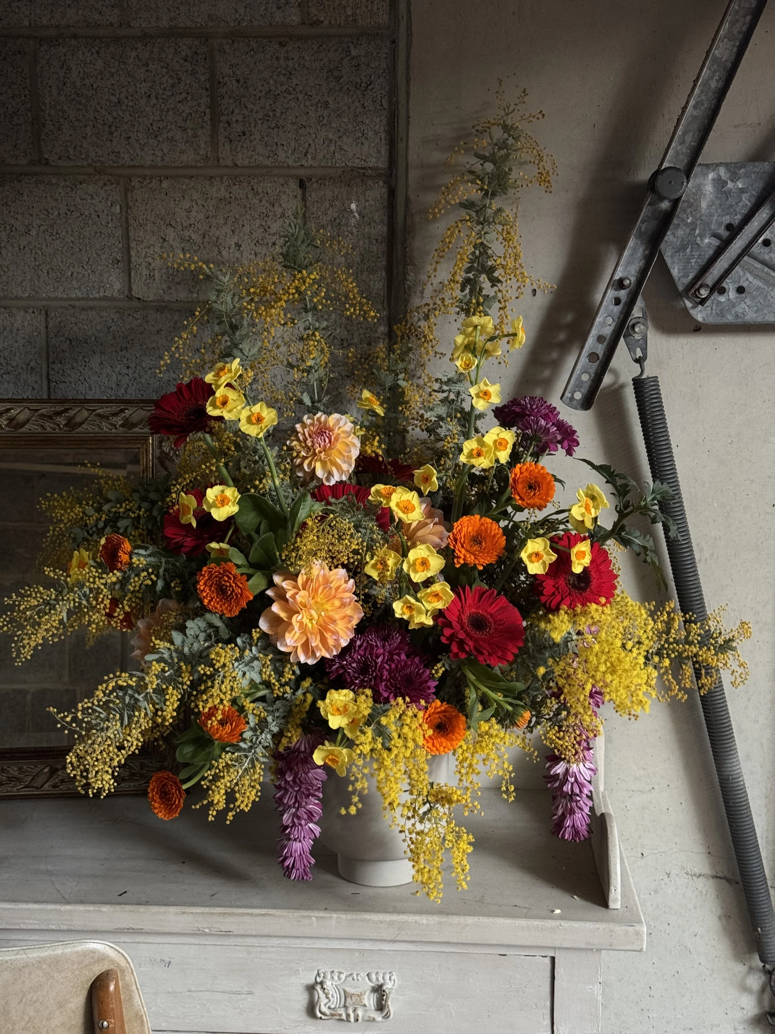 Colourful abundant large floral arrangement with yellow, orange, red, pink, and purple flowers and wattle foliage created for religious celebration