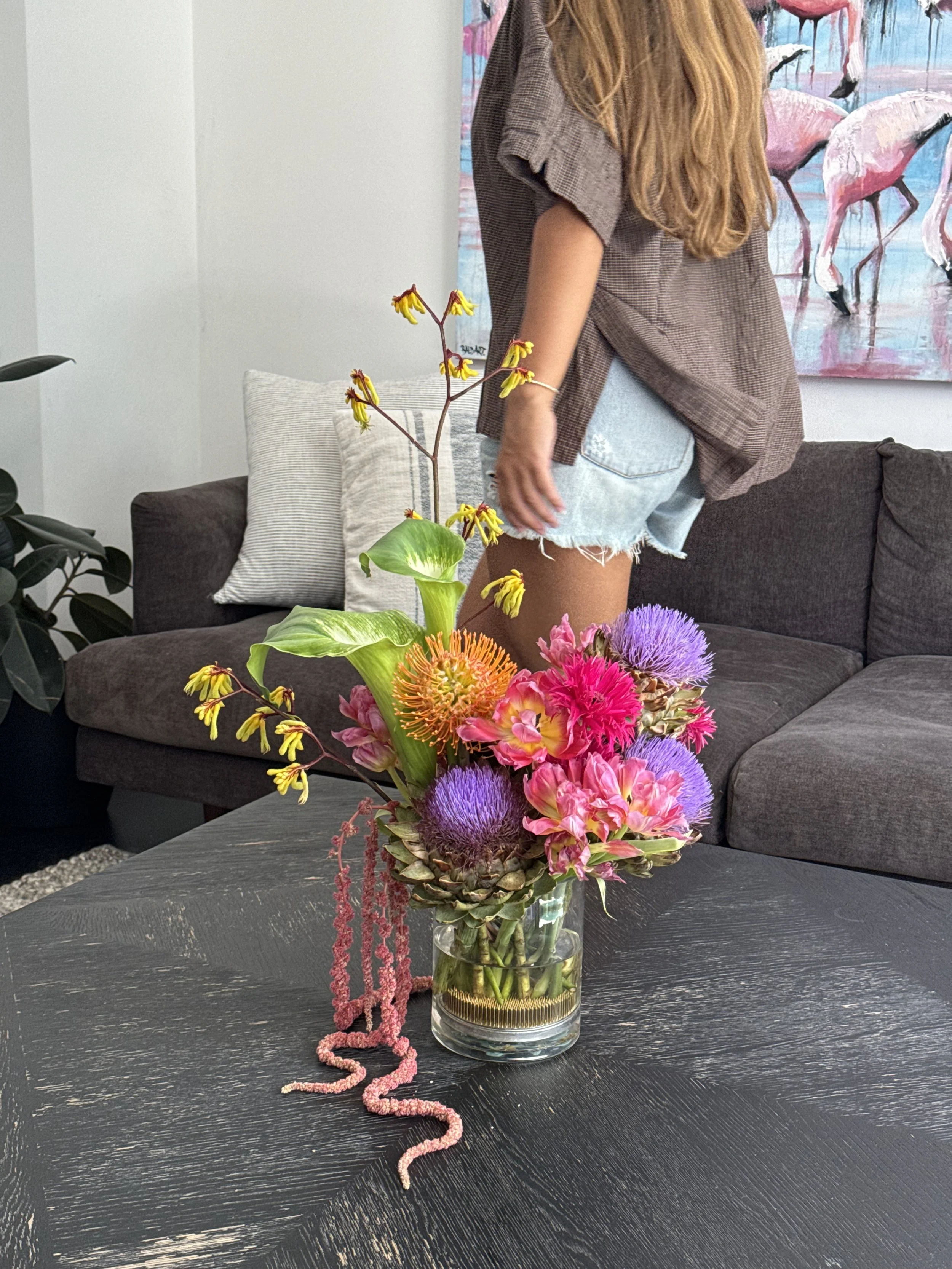 Colourful sculptural flower arrangement with pink, purple, orange flowers and green goddess' in a glass vase, on a black wooden table in a modern living room with groovy art in background. Floral arrangement styled for real estate house viewing