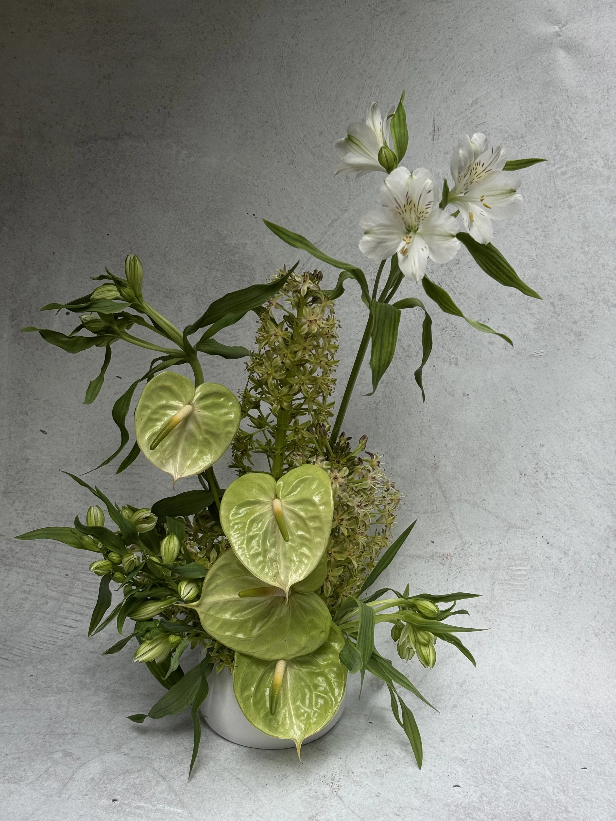 A sculptural textural flower arrangement in a white vase with white alstroemeria, green anthuriums, and pineapple lilies against a concrete backdrop.