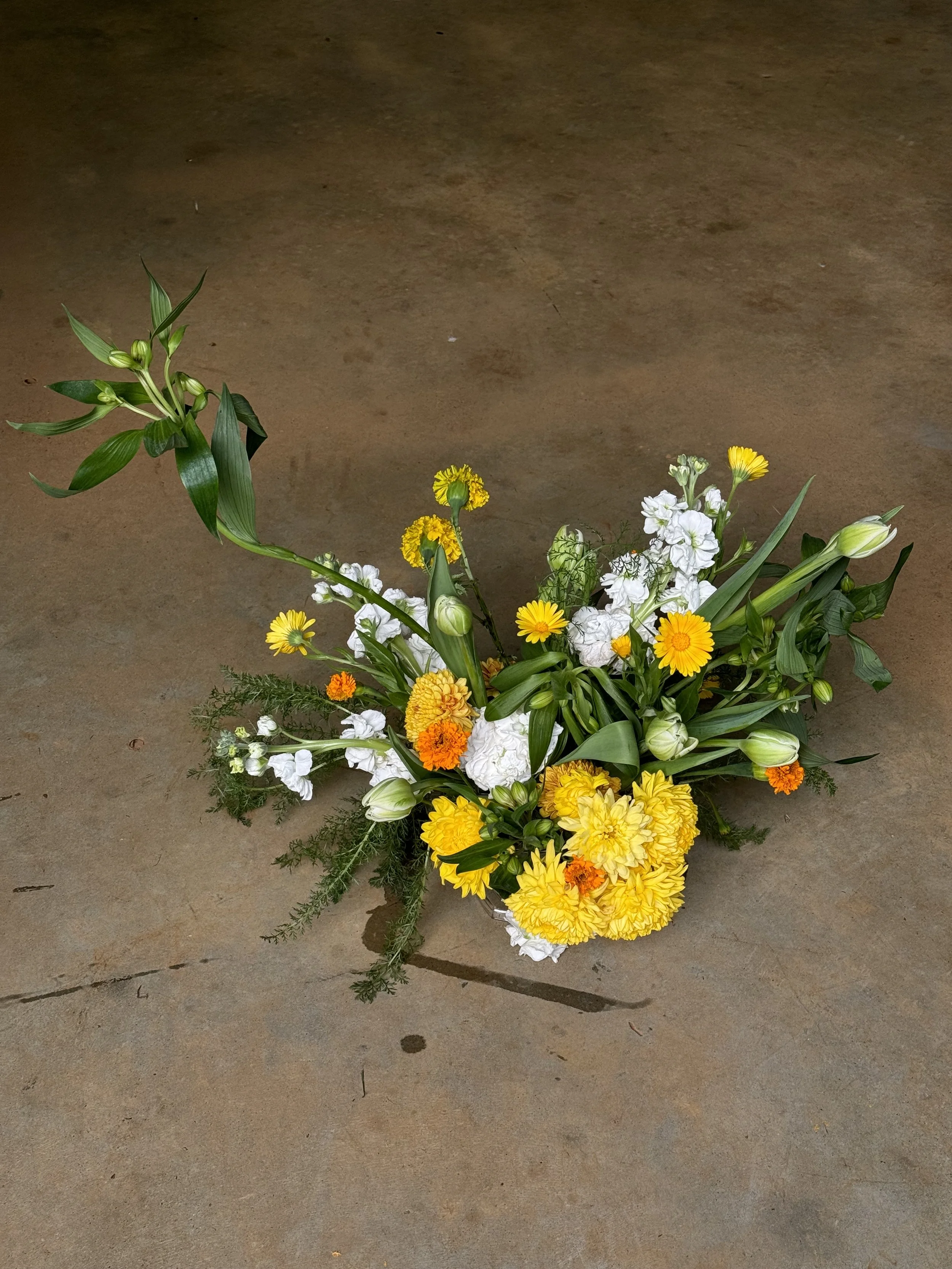 A flower arrangement on a concrete floor featuring yellow, white, and orange flowers and green foliage.