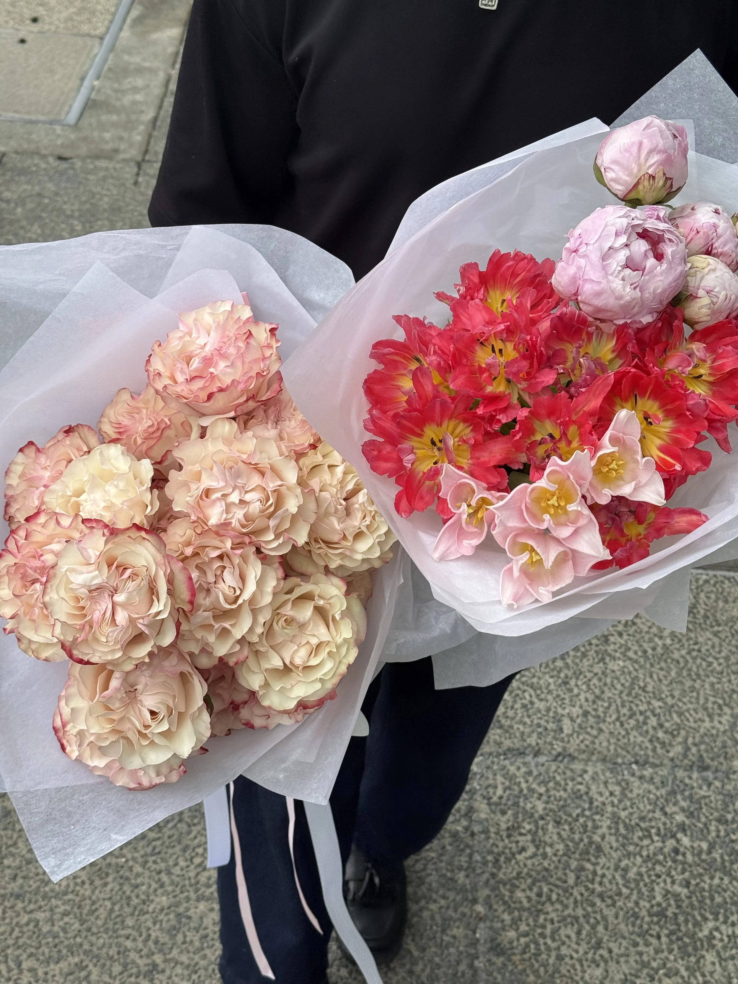 A person holding two bouquets of pink and white flowers wrapped in white paper, standing on a sidewalk.