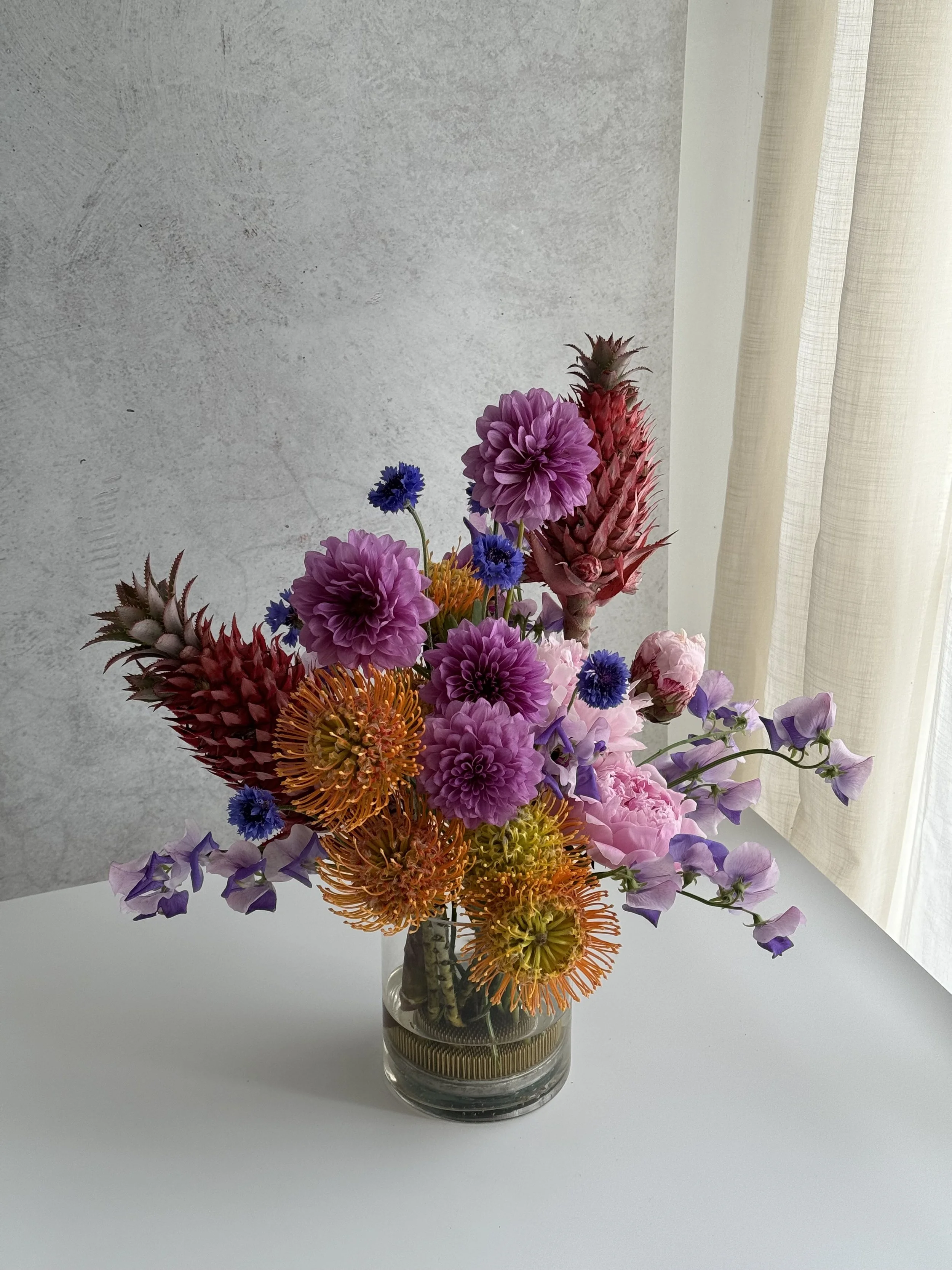 Colourful floral arrangement in a glass vase on a white table near a window with curtains. Pink pineapples, peonies, sweet pea included in design.