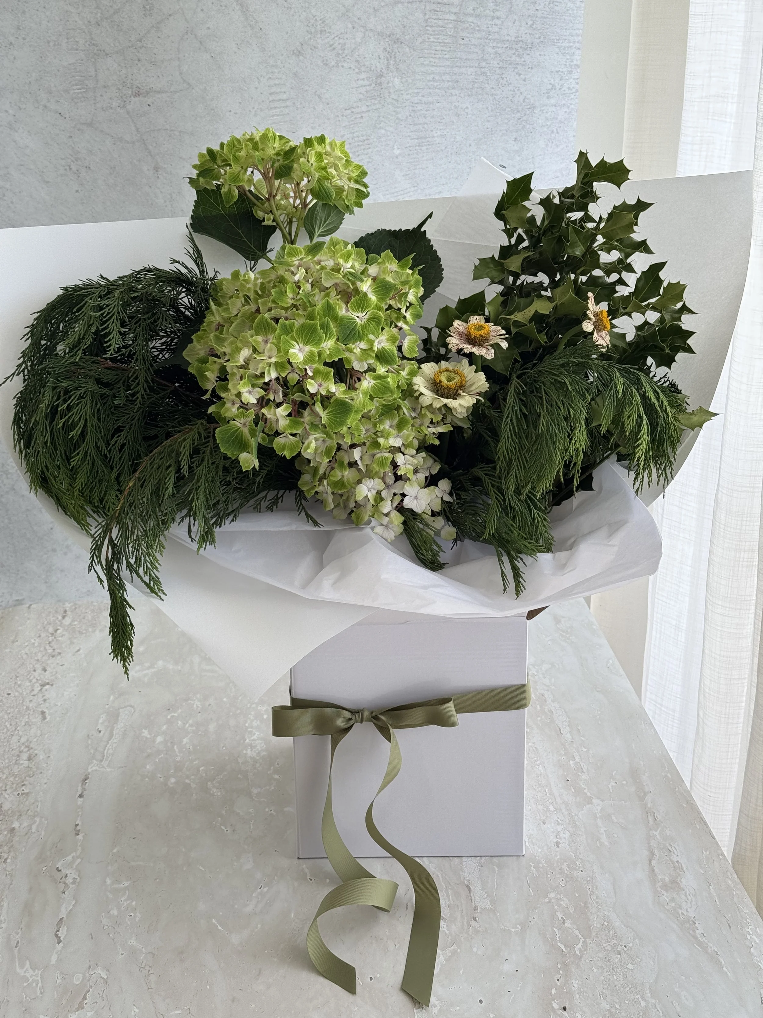 A flower arrangement in a white gift box with a green bow, containing green hydrangeas, holly leaves, sprigs of pine, and white zinnias, wrapped in white tissue paper. Christmas flowers. gifted delivery