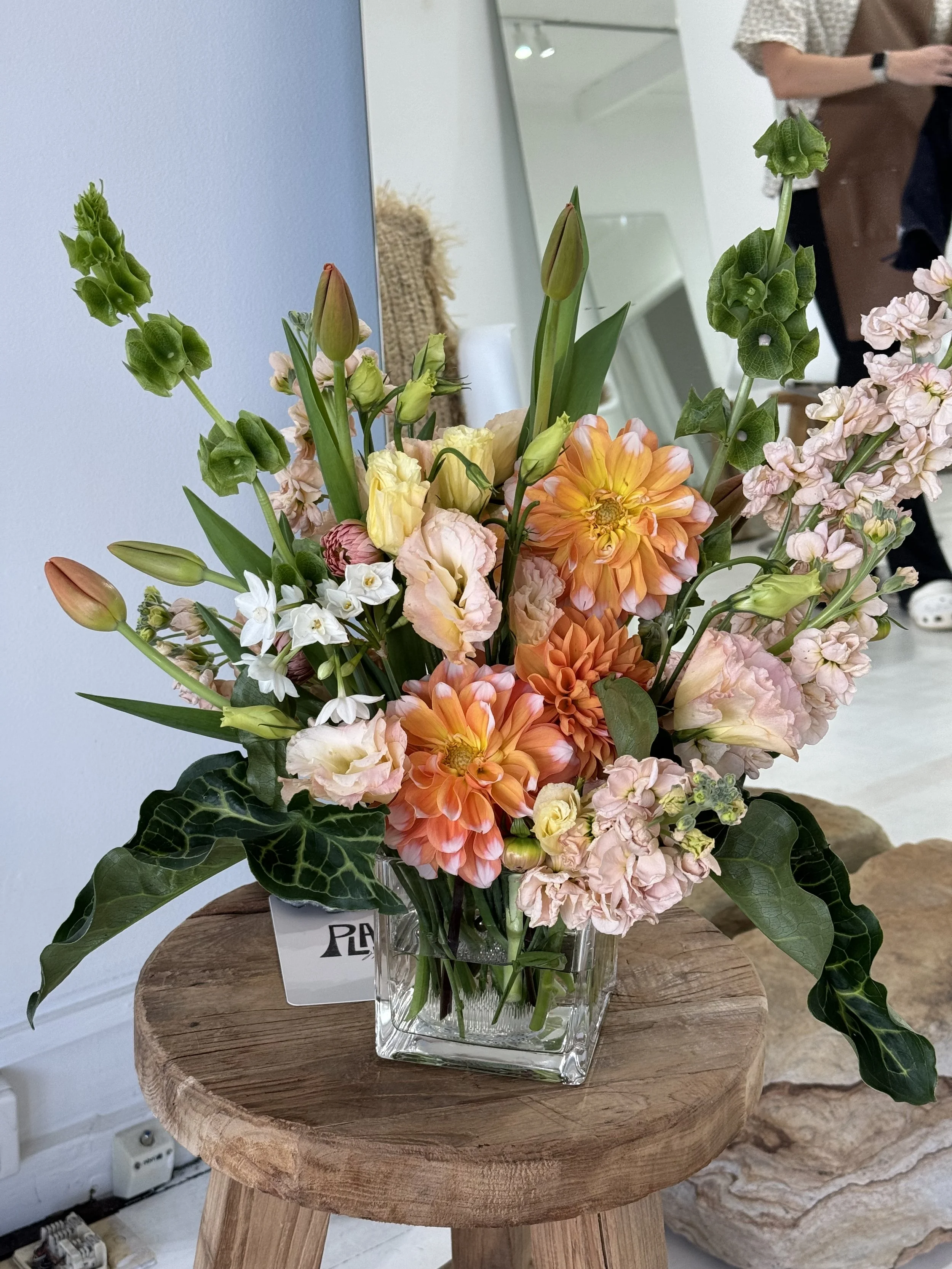 A custom floral arrangement of various peach and green flowers in a glass vase on a wooden stool inside/styled for a hair salon