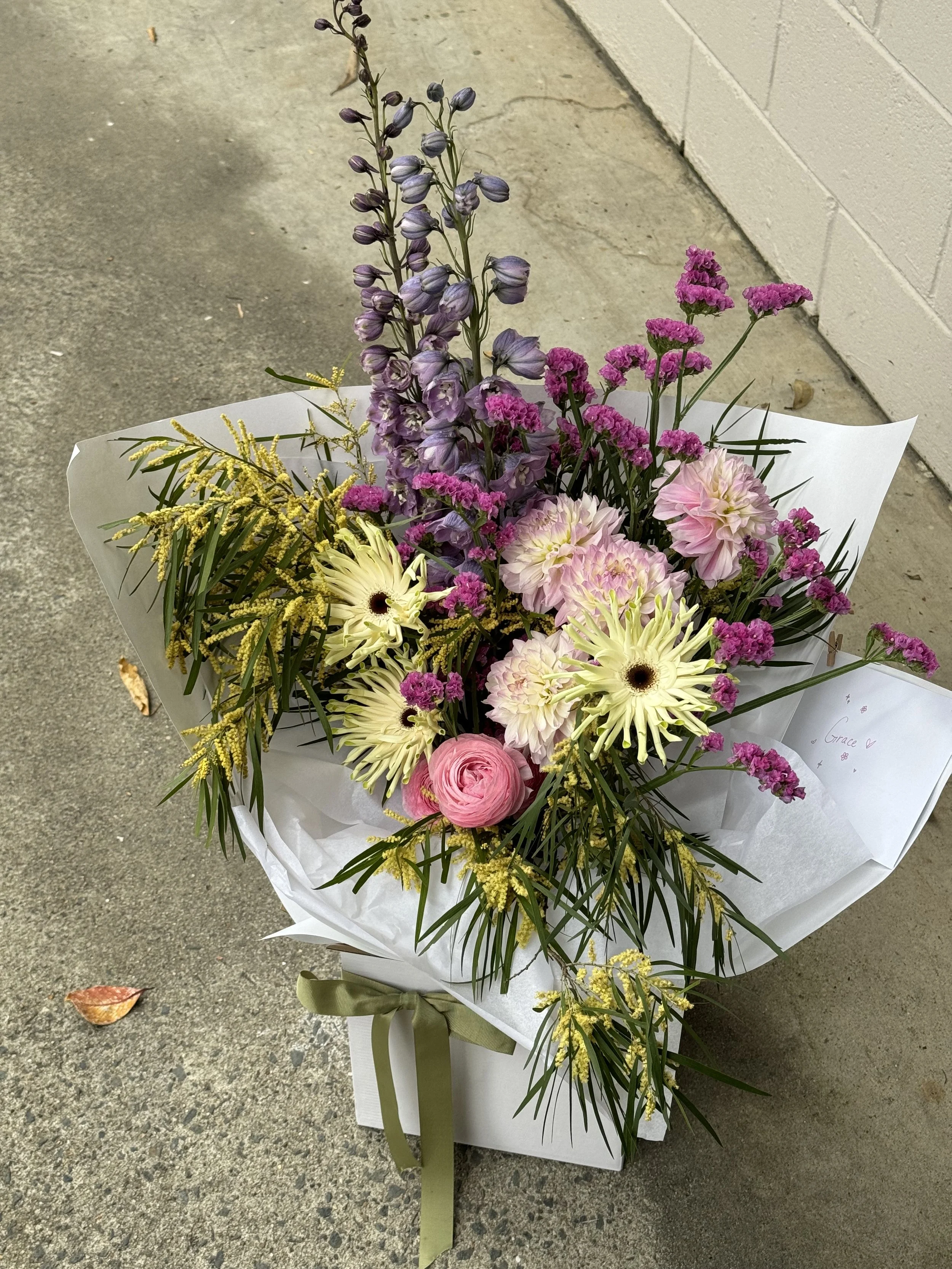 A gifted flower bouquet of mixed pink, purple, yellow flowers arranged in a white gift box and wrapping with a green bow.
