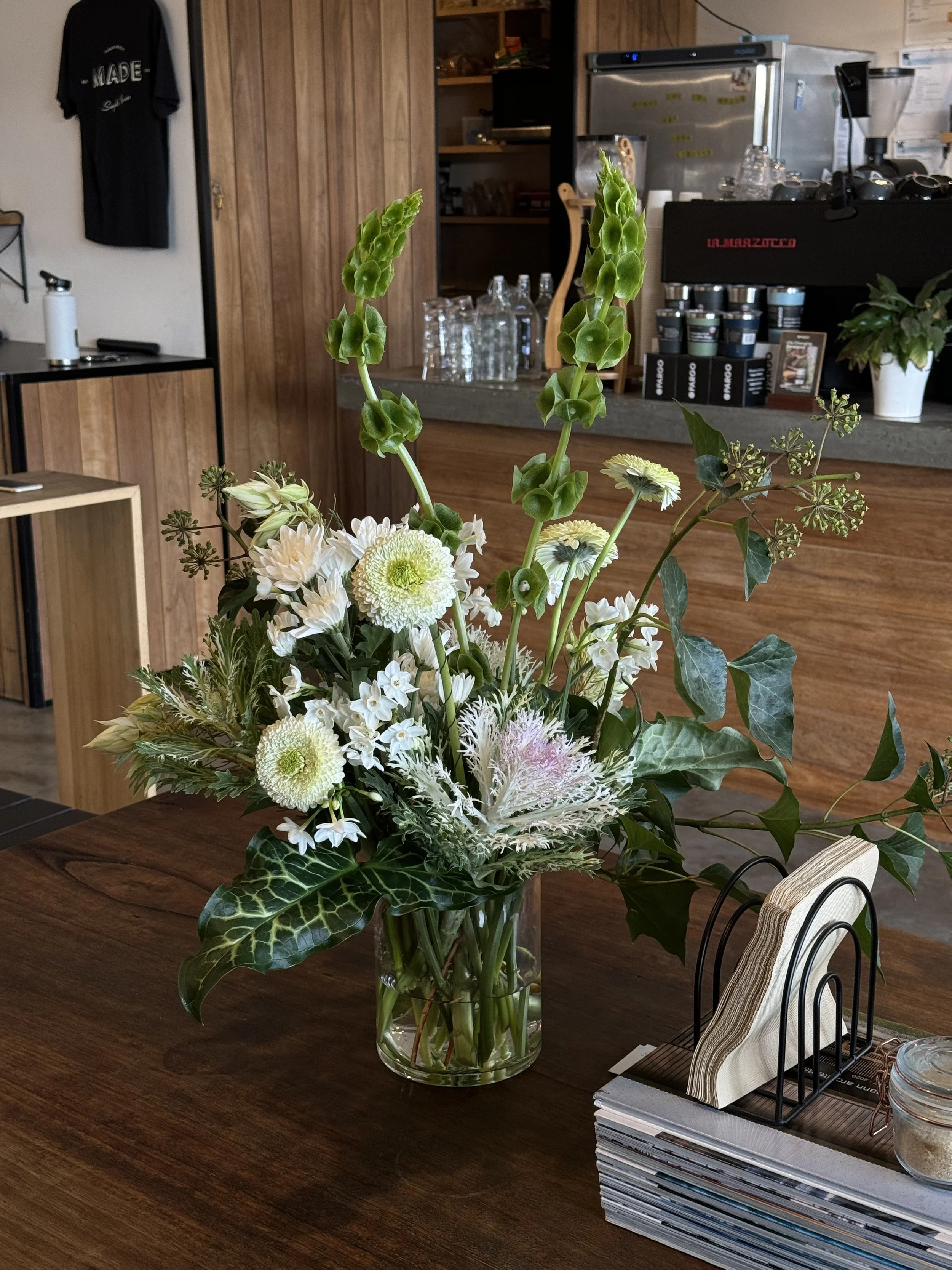 A bouquet of white flowers with green foliage in a clear glass vase on a wooden table inside a cafe 