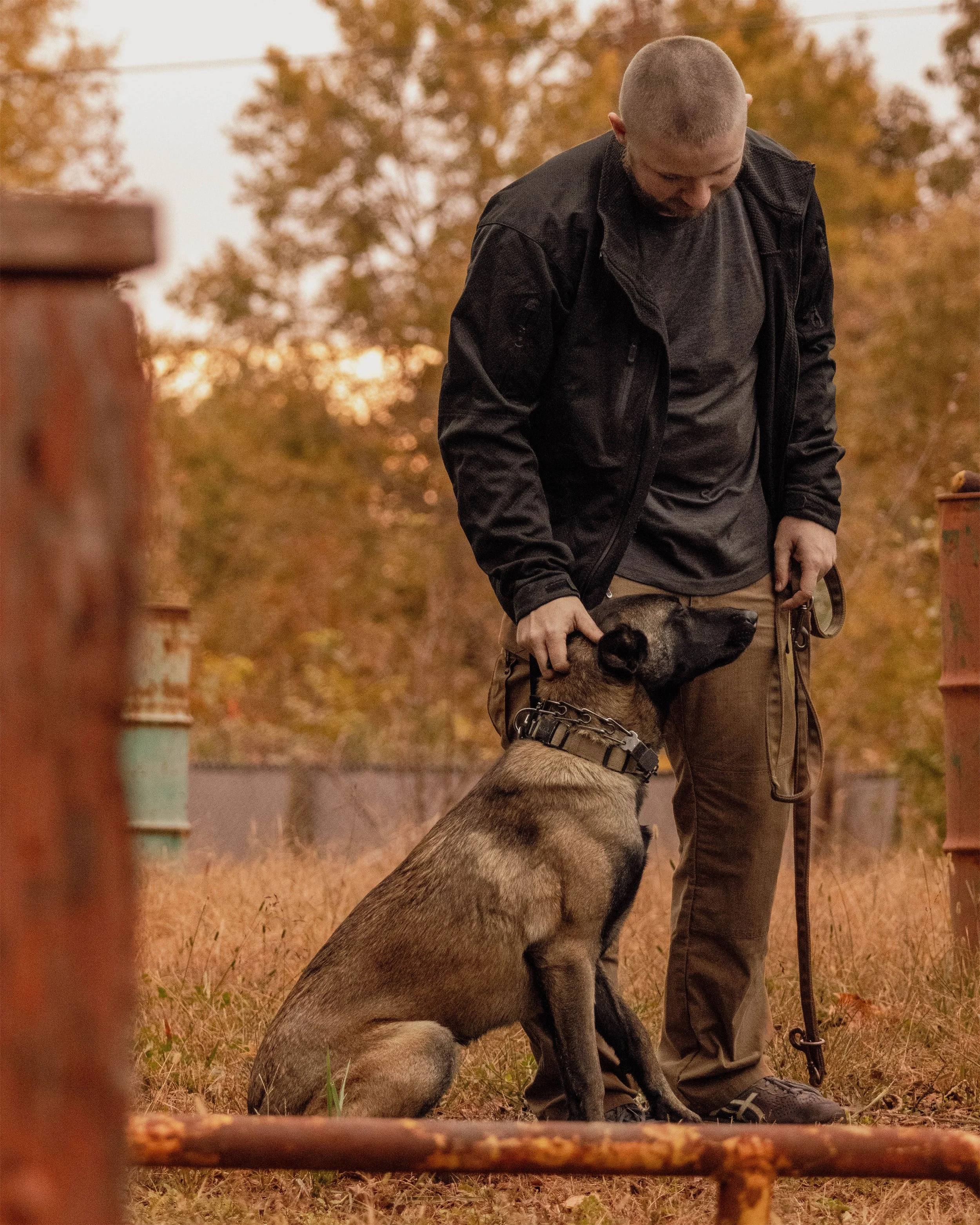 A man in black jacket and gray shirt standing outdoors with a large dog sitting next to him during fall, with orange and brown trees in the background.