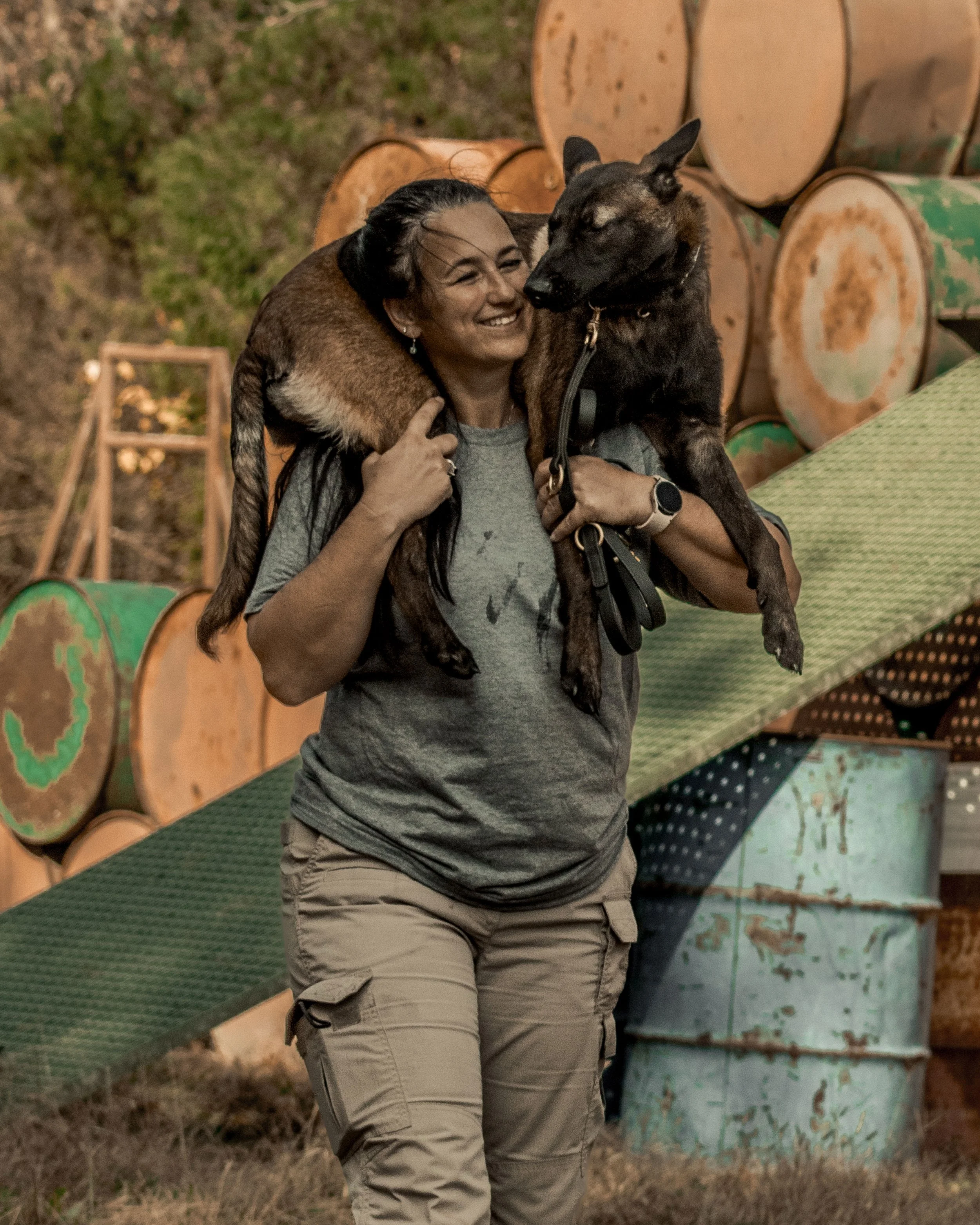 A woman smiling while holding two dogs, one on her shoulders and one in her arms, in an outdoor setting with stacked logs and barrels in the background.