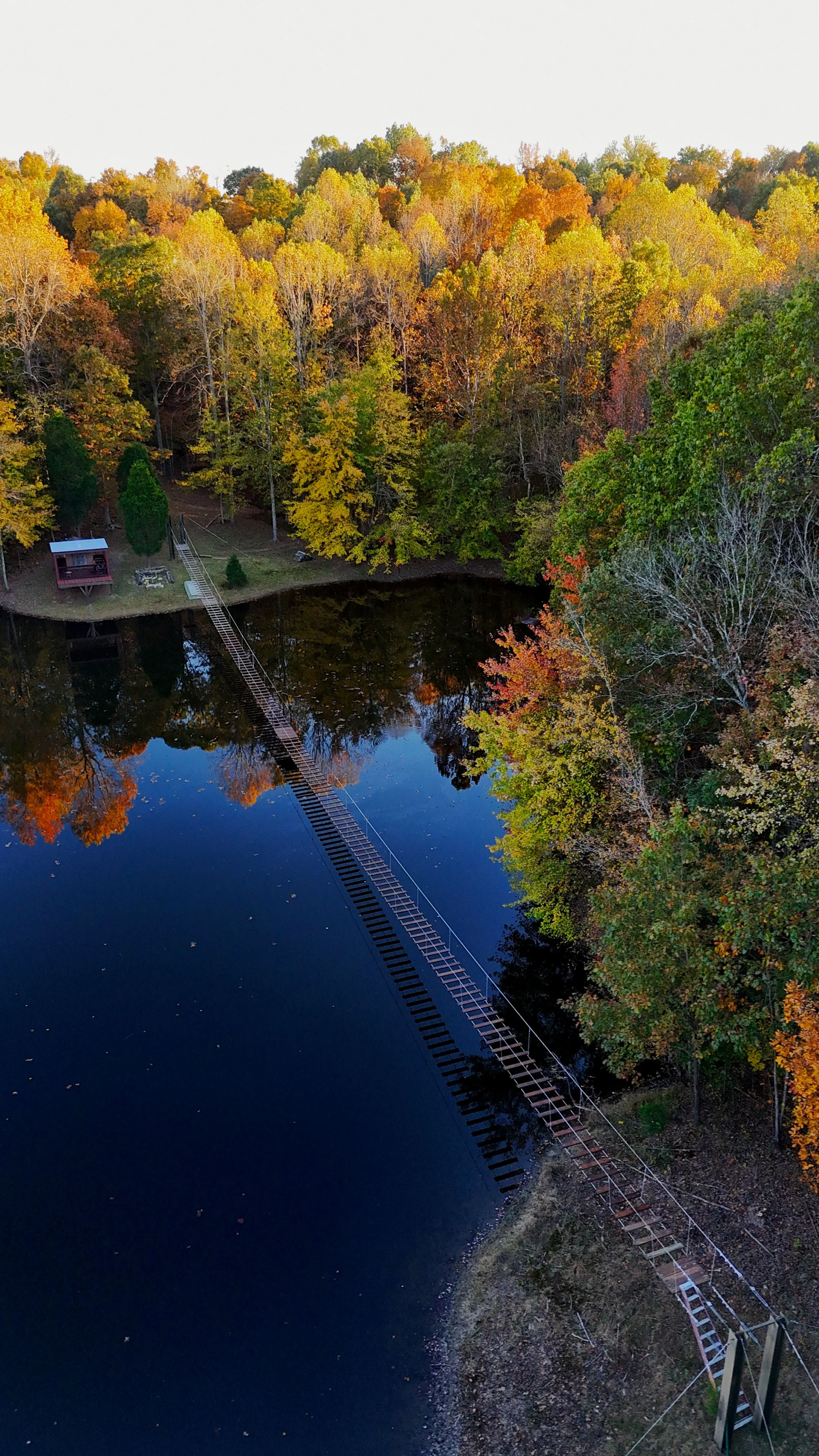 Aerial view of a river surrounded by autumn trees with yellow, orange, and green leaves, and a suspension bridge crossing over the river.