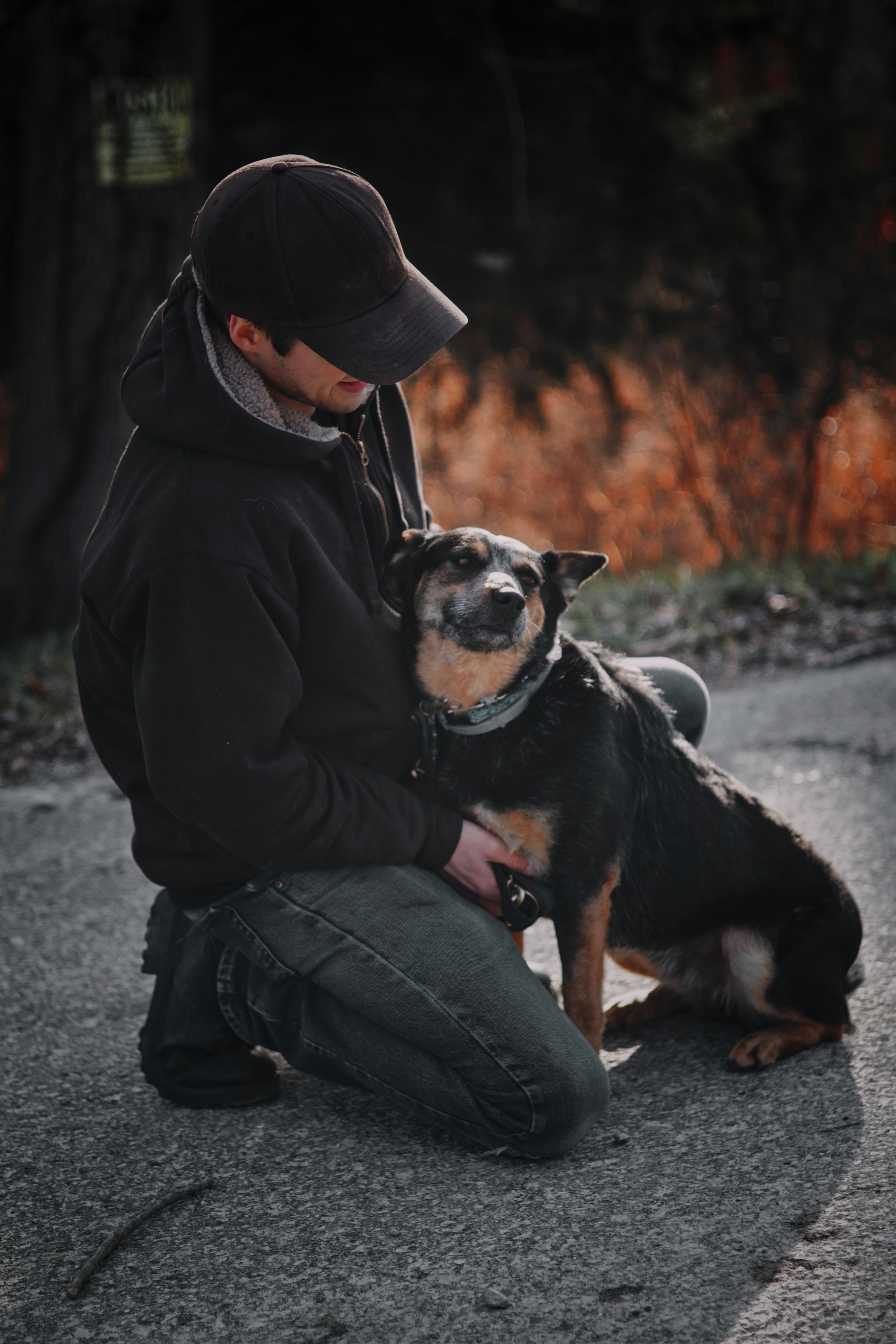 A person wearing a black hoodie and cap kneeling on the ground, hugging a black and tan dog outdoors at dusk with fall foliage in the background.