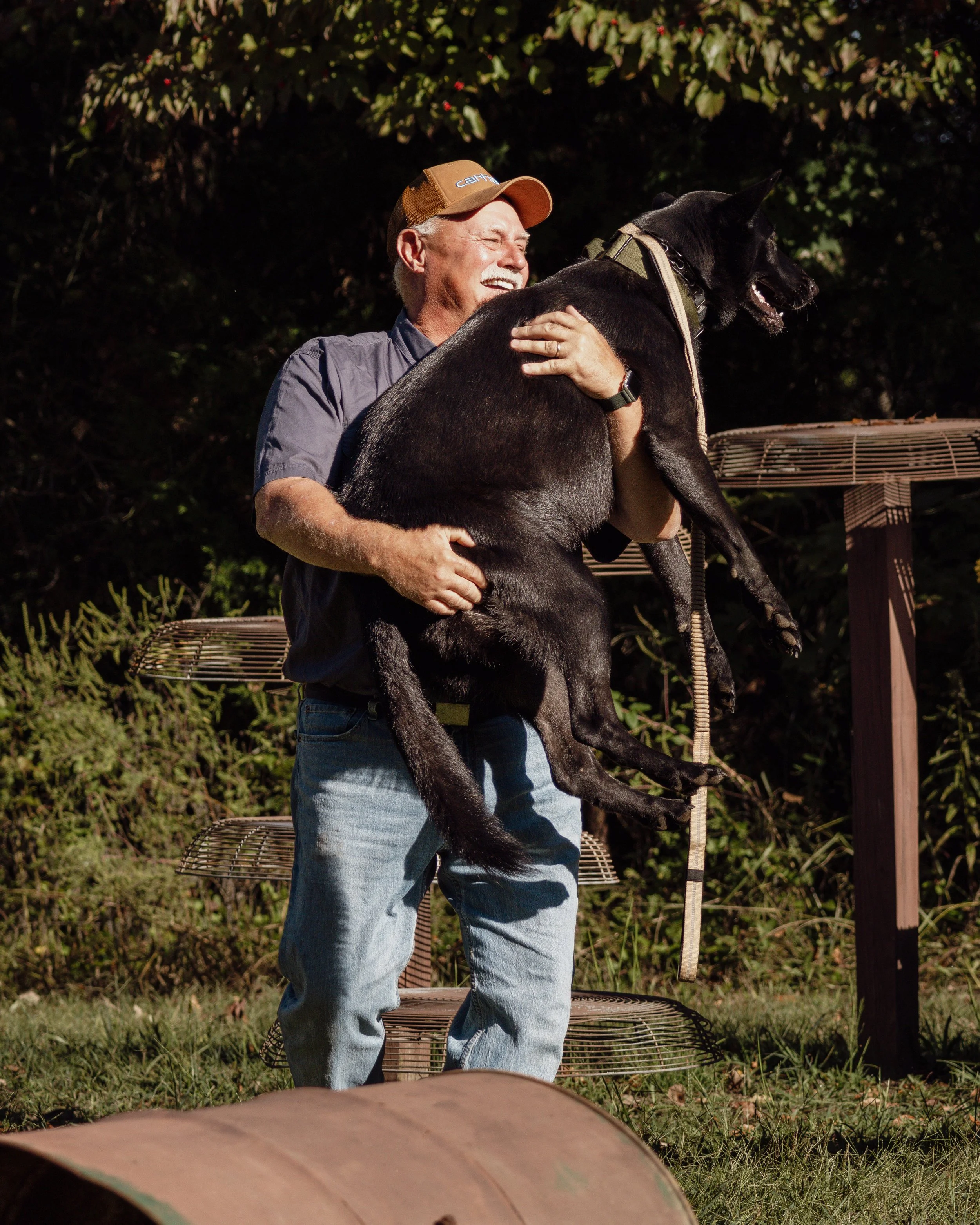 A smiling man lifting a black dog outdoors during daytime with greenery in the background.