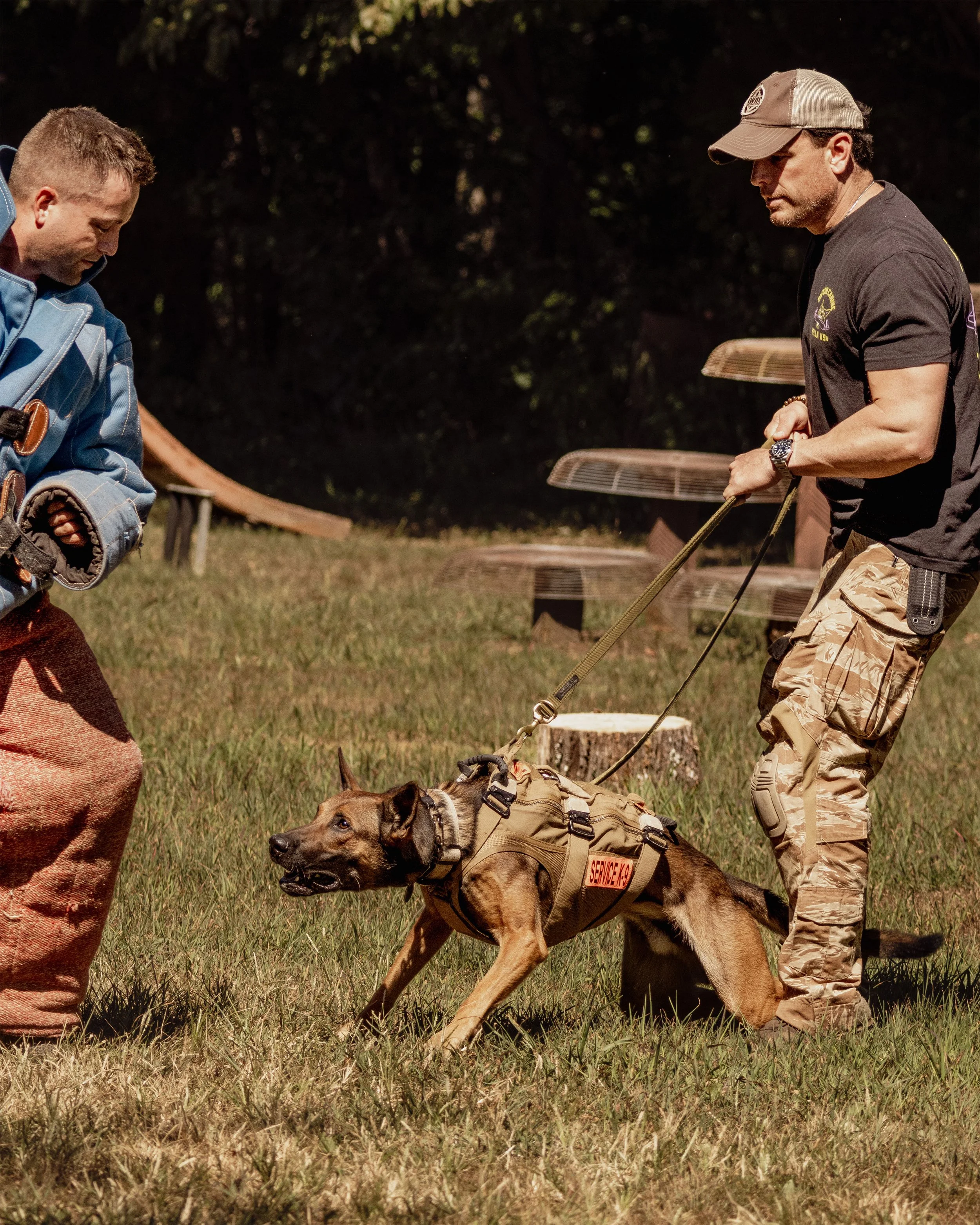 A man in camo pants and a black shirt holding a service dog on a leash, with another man in a blue jacket nearby in an outdoor setting.