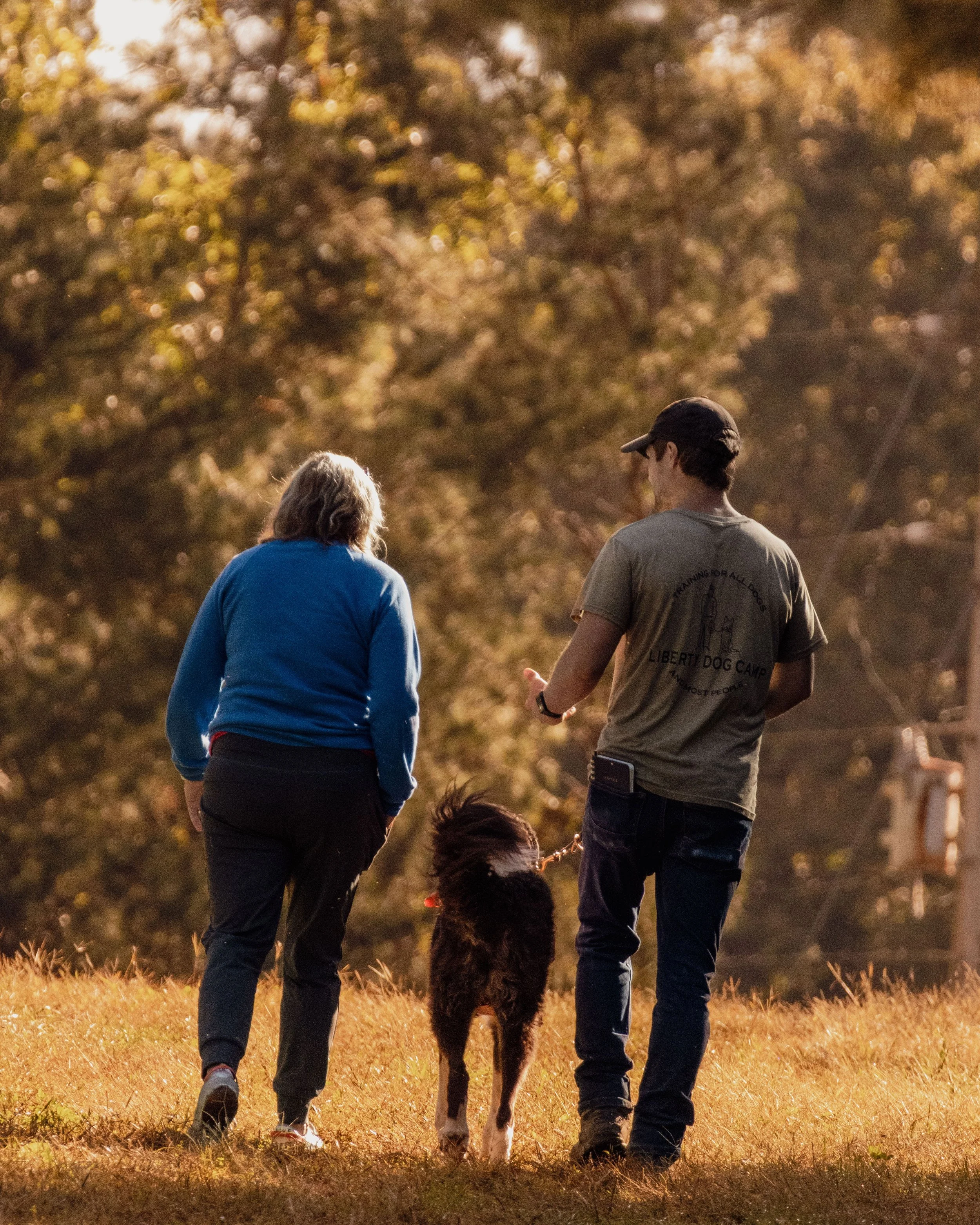 Two people walking a dog in a park during fall, with trees in the background.