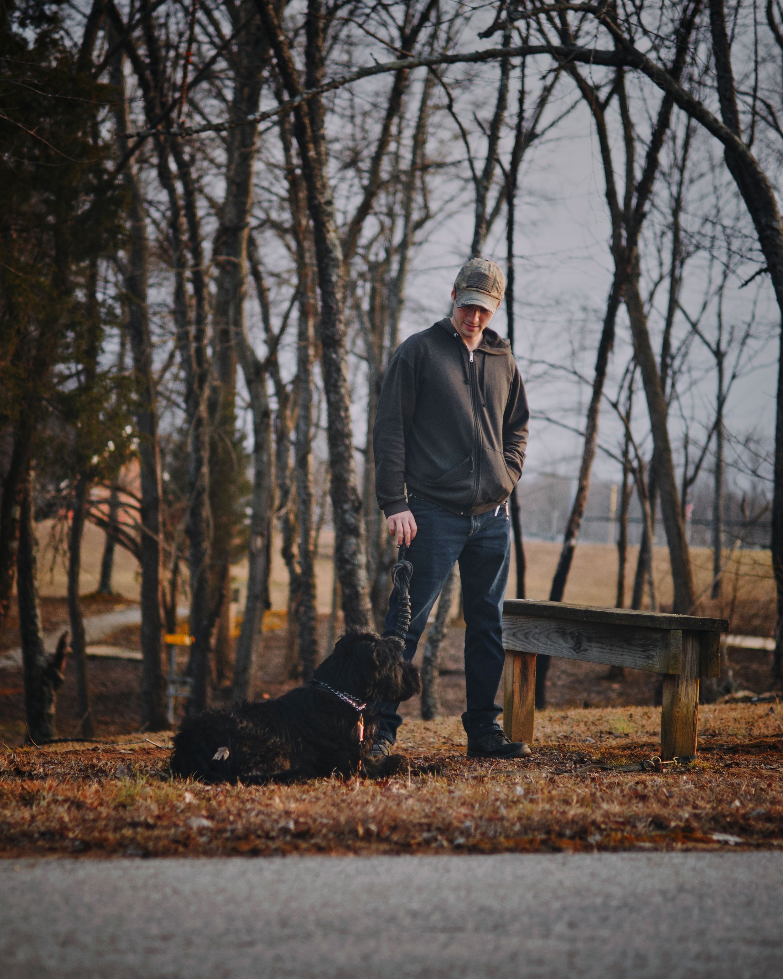A man holding a leash attached to a black dog sitting on the ground, outdoors in a wooded area during late afternoon or early evening.