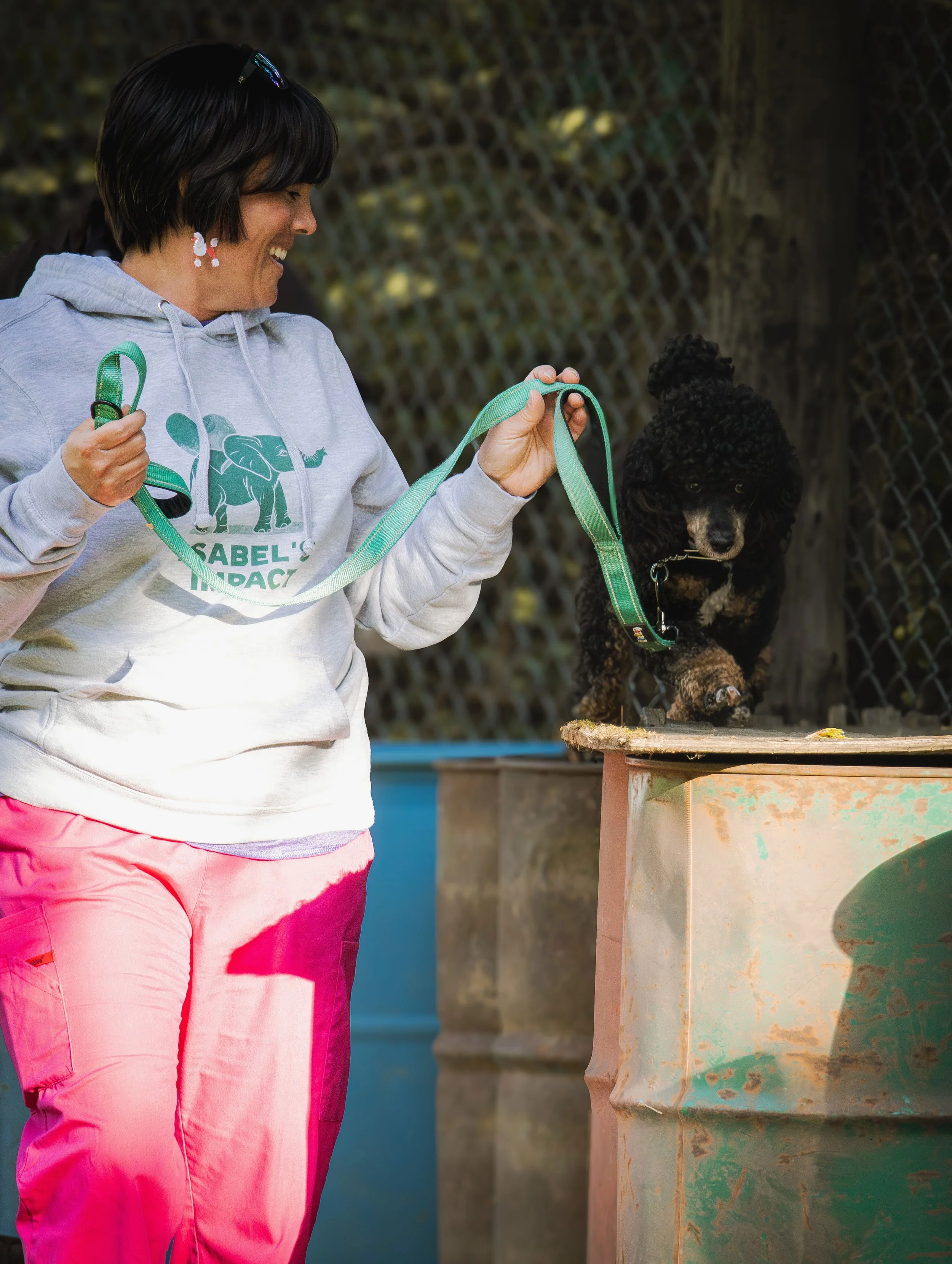 A woman smiling and holding a green leash with a black poodle standing on a raised surface outdoors near a chain-link fence.